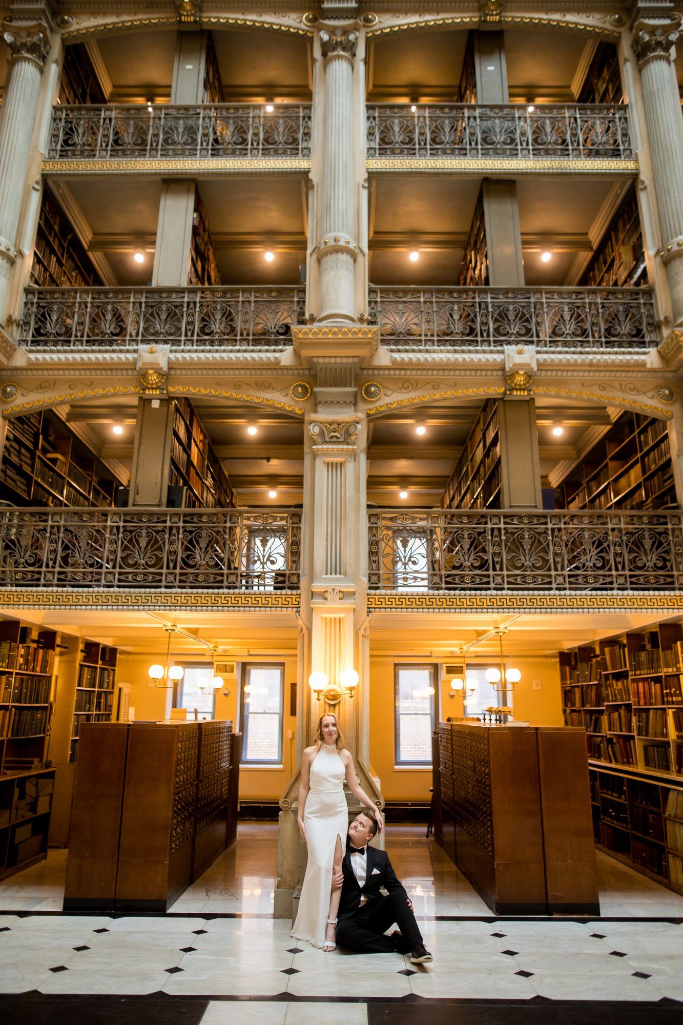 Bride and groom posing in a grand multi-story library with ornate iron balconies and floor-to-ceiling bookshelves.