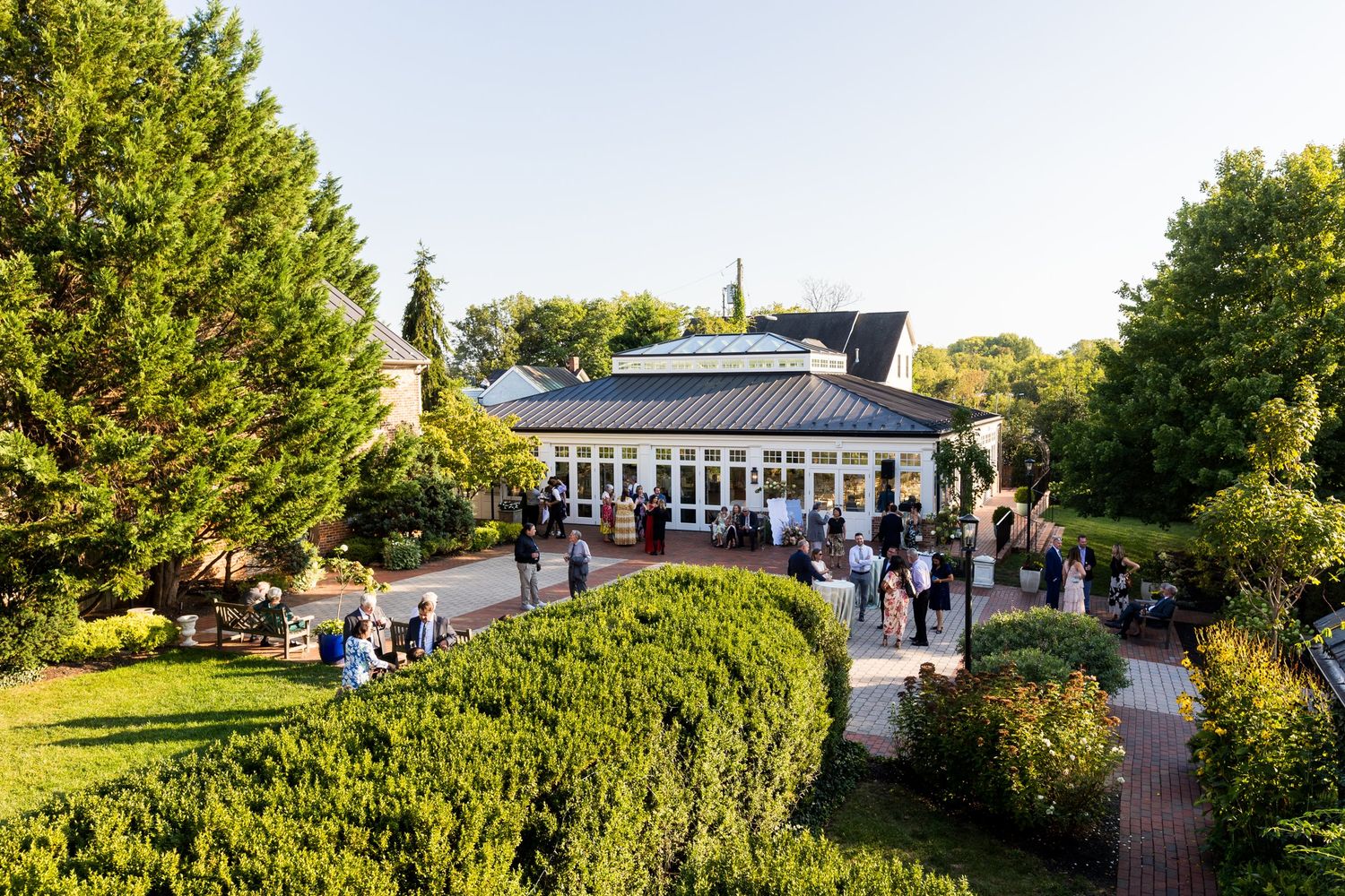 Elegant garden venue with lush greenery and a glass conservatory building surrounded by guests at an outdoor event.