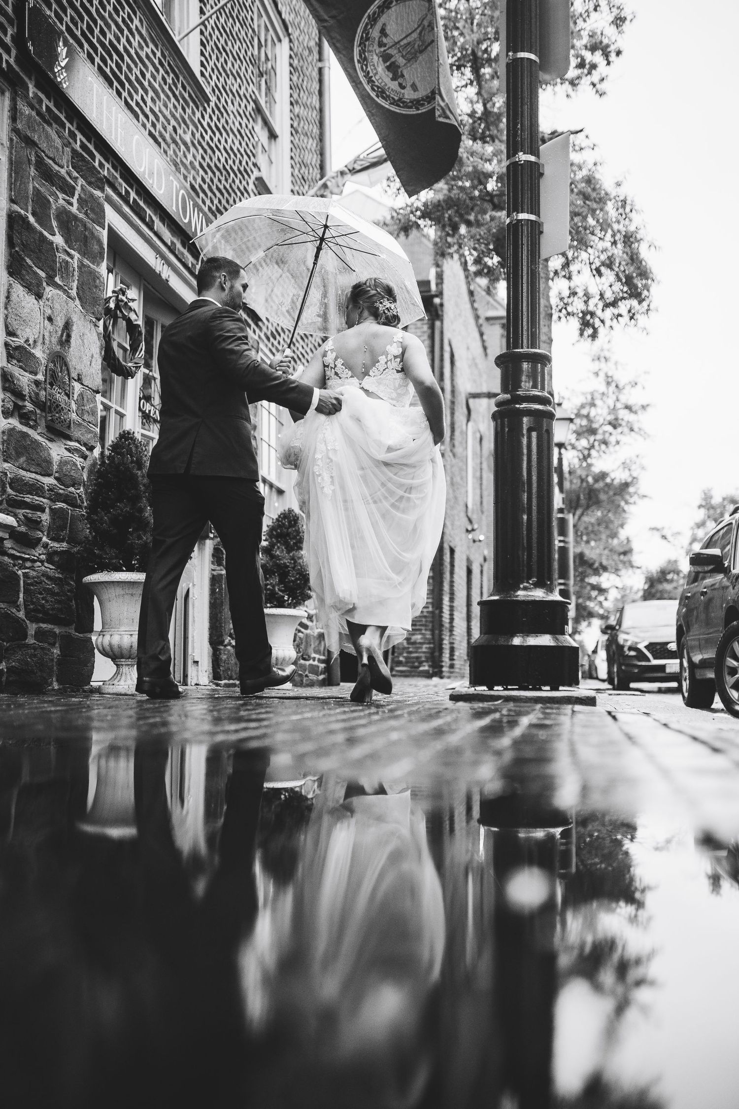 Black and white photo of bride and groom walking under umbrella on rainy cobblestone street with puddle reflection.