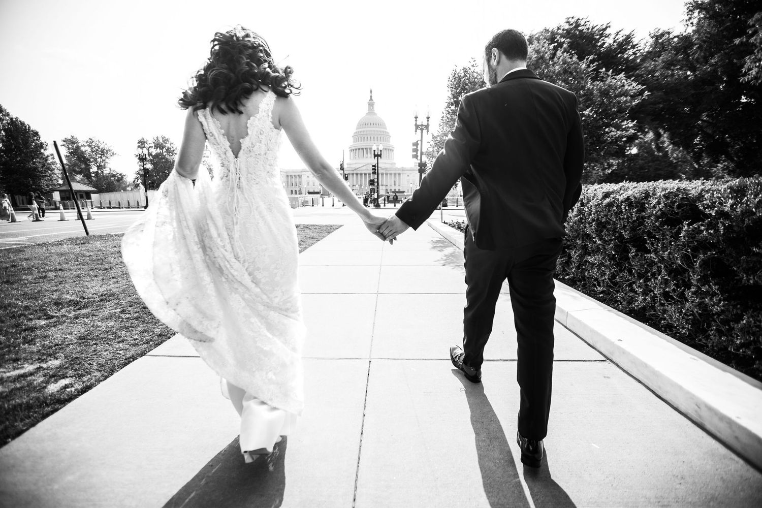 Black and white photo of a bride and groom holding hands walking toward the US Capitol building on their wedding day.