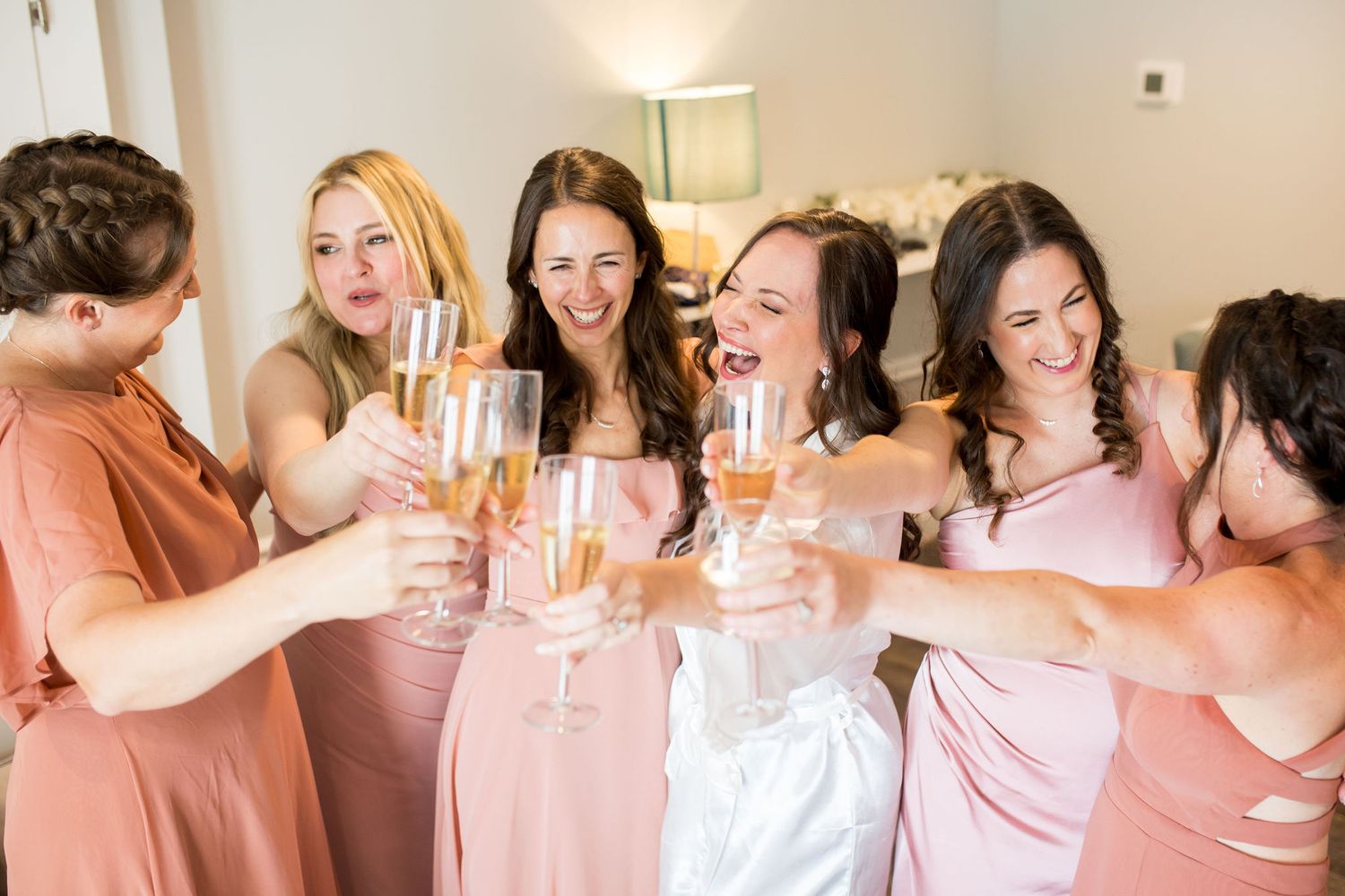 Bride and bridesmaids in pink dresses toasting with champagne glasses during wedding day preparations.
