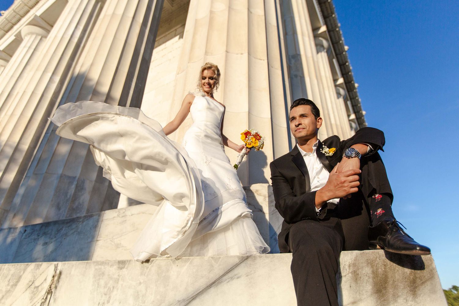 Bride in flowing white gown holding bouquet stands beside groom in black suit on marble steps of elegant columned building.