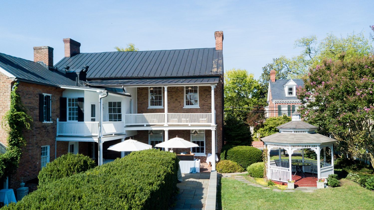 Historic brick manor with black metal roof, white gazebo, outdoor dining area, and manicured gardens on a sunny day.