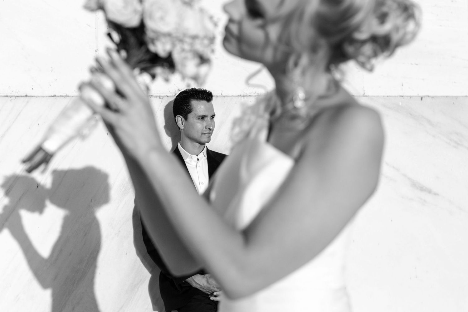 Black and white wedding photo of groom gazing at bride holding bouquet, with shadow cast on white wall behind them.