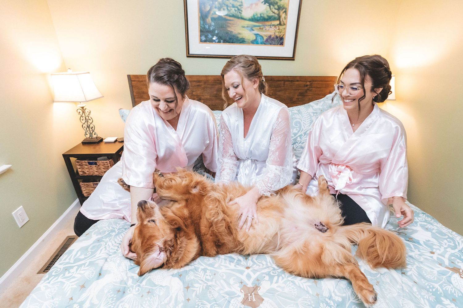 Three bridesmaids in pink robes laughing with two golden retrievers on a bed during wedding preparations.