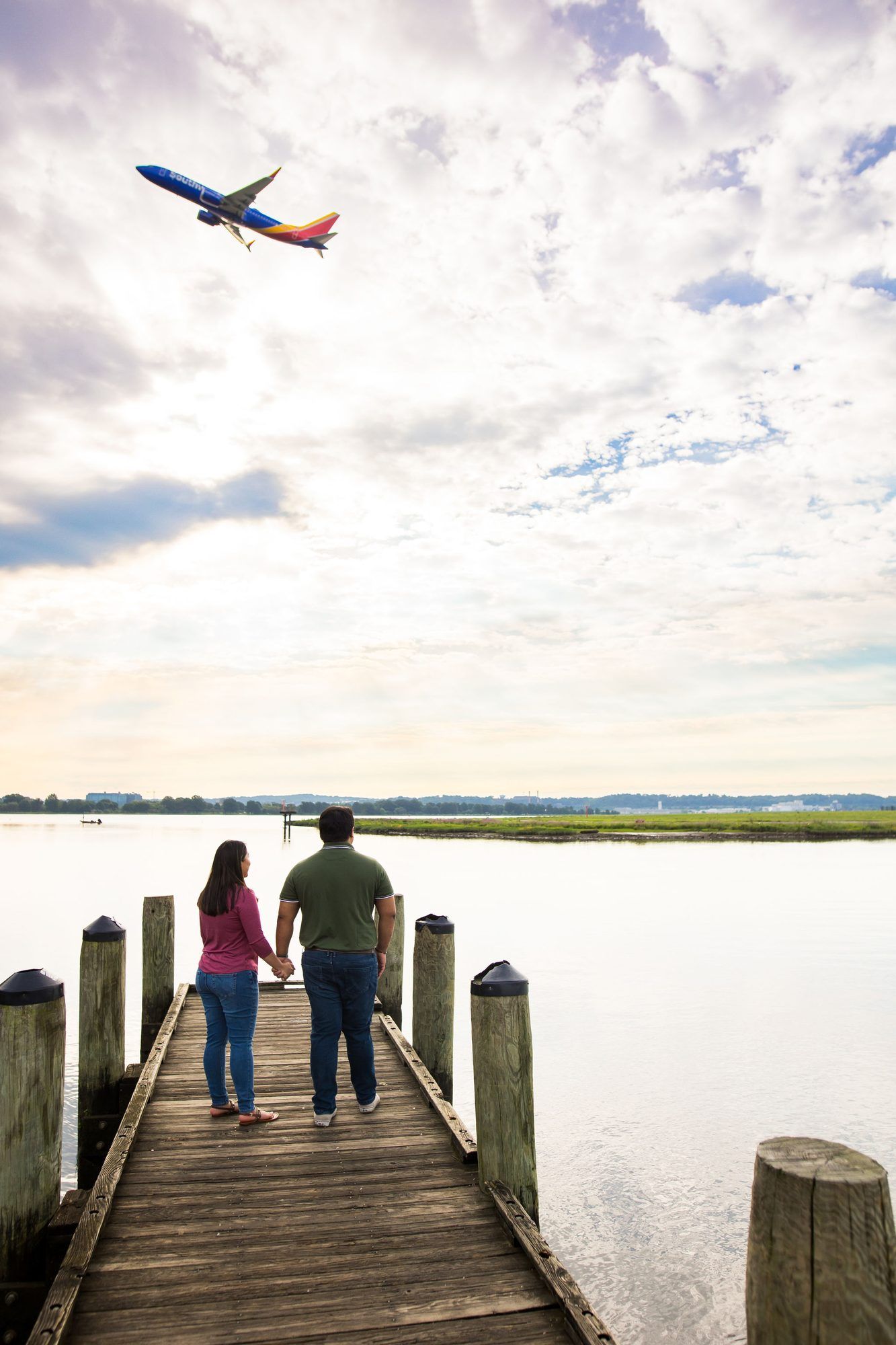 Couple holding hands on wooden dock overlooking calm water as Southwest Airlines plane flies overhead under cloudy sky.