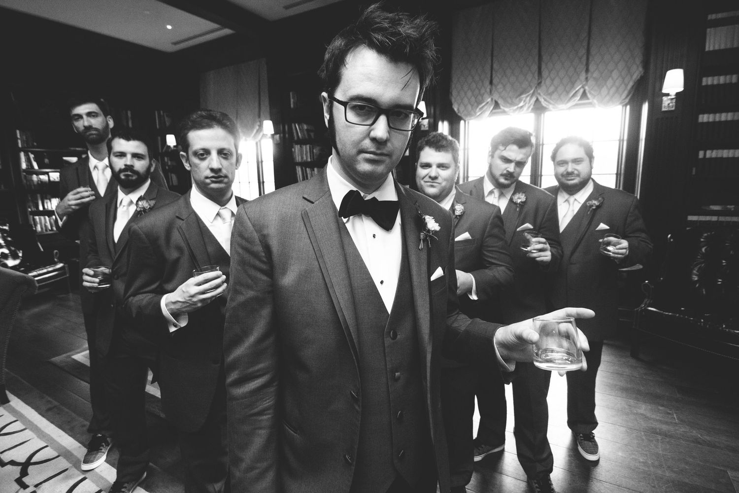 Groom in bow tie suit poses with groomsmen holding drinks in black and white wedding photo.