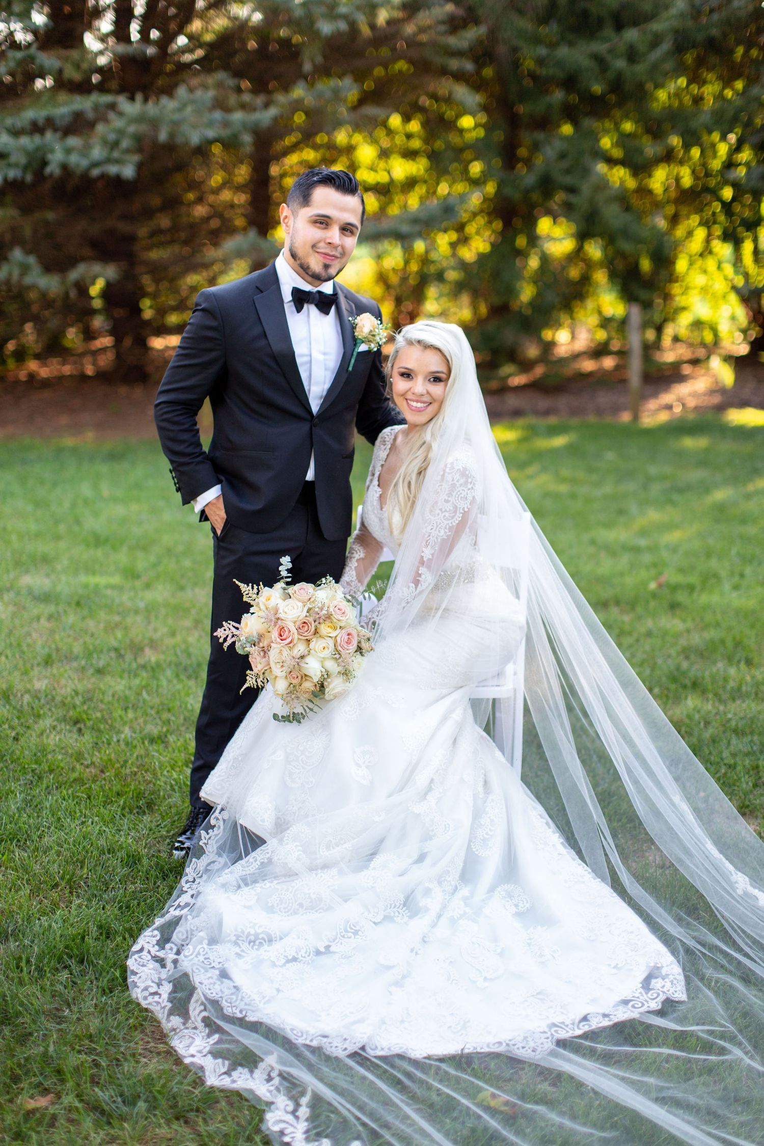 Bride in lace gown with cathedral veil holds bouquet beside groom in black tuxedo on green lawn.