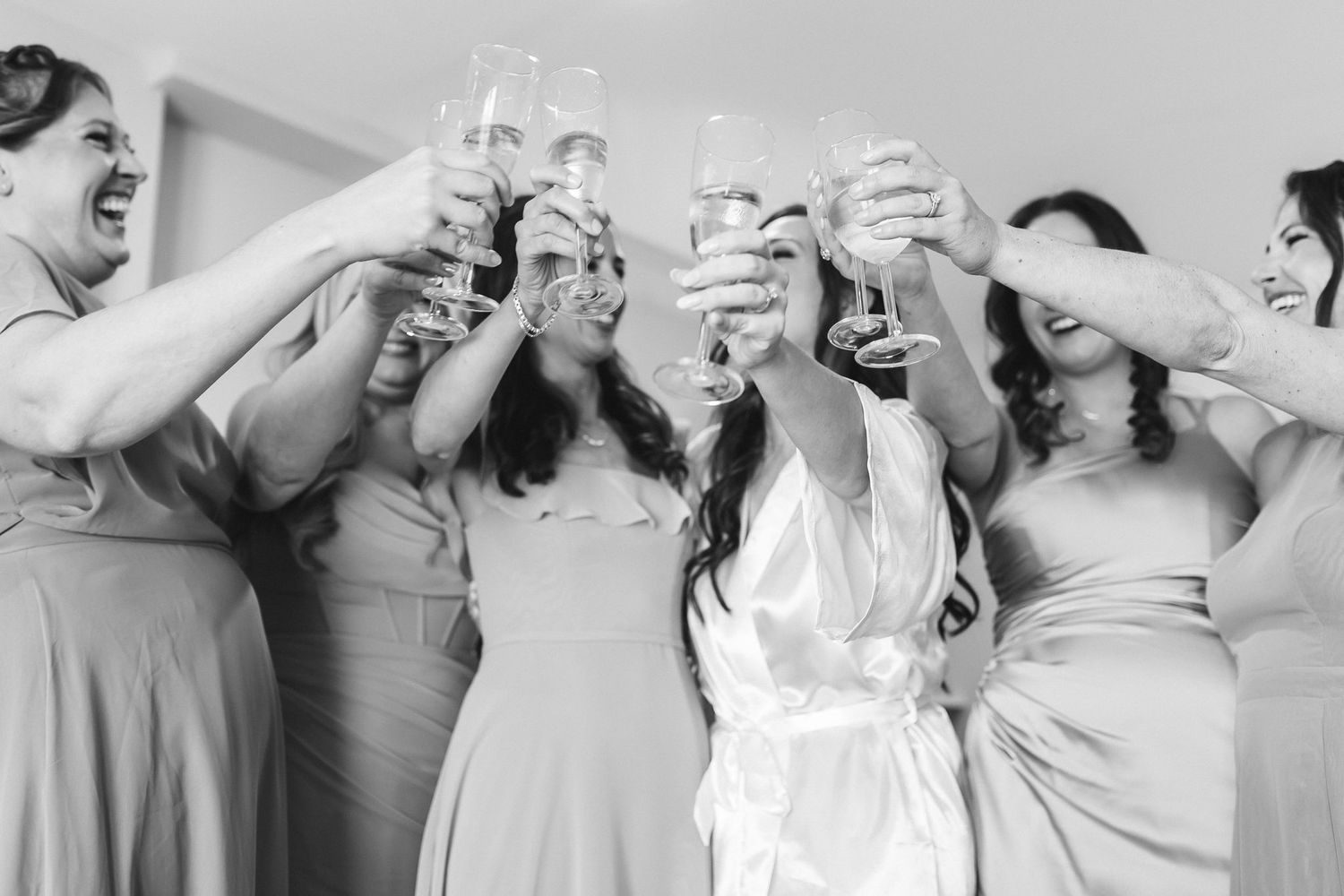 Bridesmaids and bride in white robe toasting with champagne glasses in a joyful black and white wedding photo.