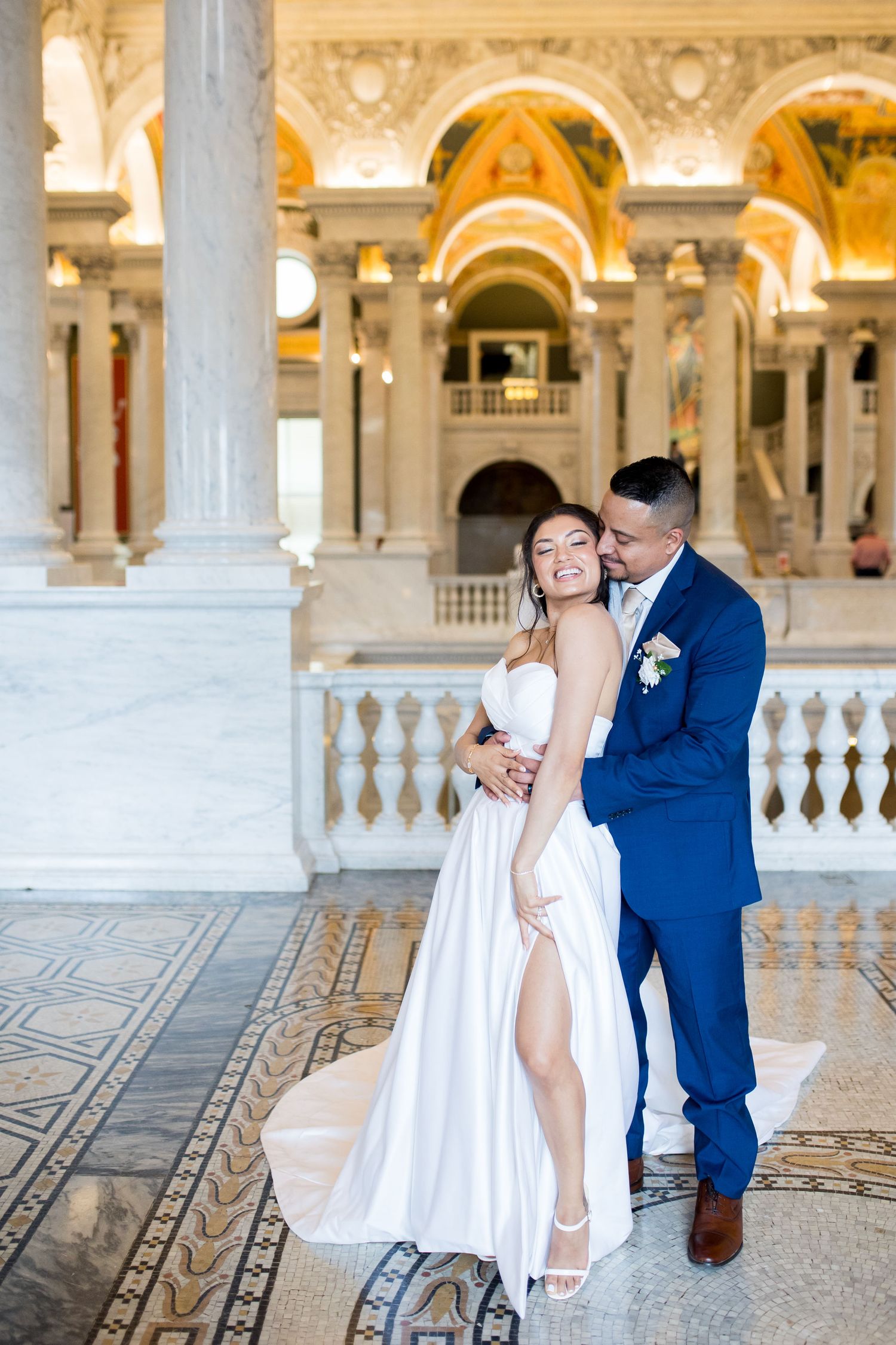 Bride and groom embrace in the elegant marble hall with ornate columns and golden ceiling at the Library of Congress.