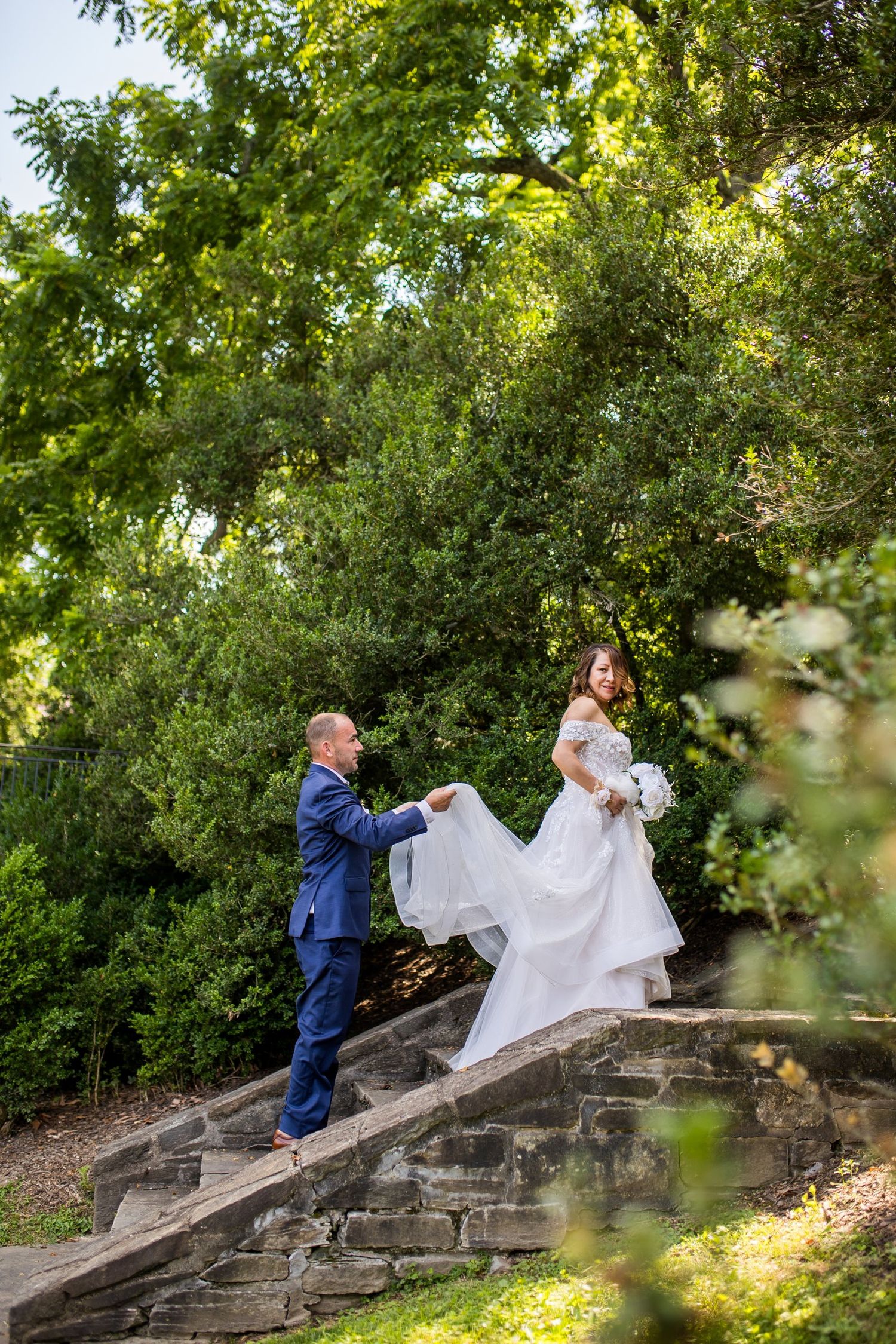 Groom in blue suit helps bride in white gown climb stone steps surrounded by lush green trees at outdoor wedding.