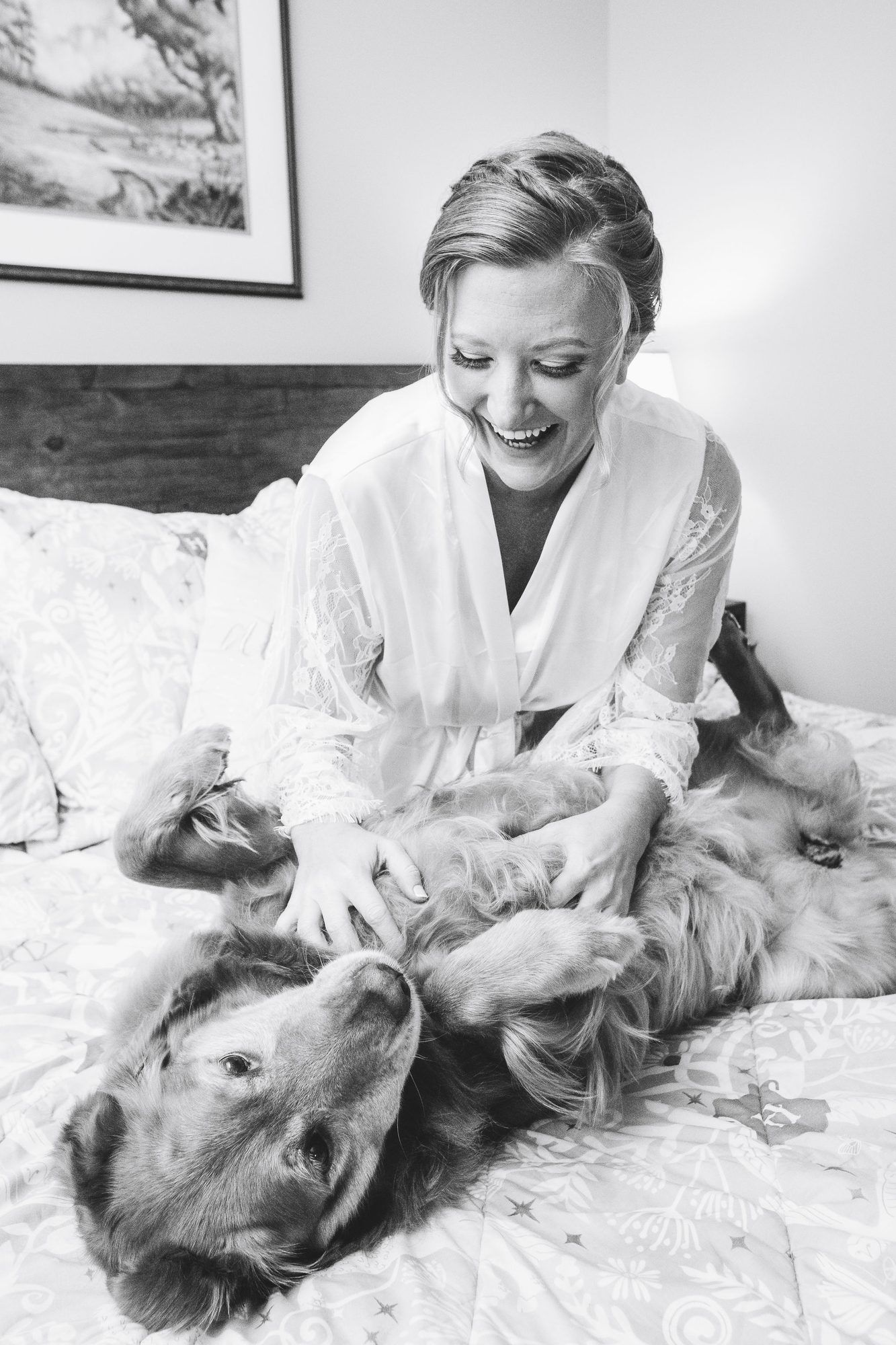 Black and white photo of a smiling bride in a white robe playing with her golden retriever dog on a bed.