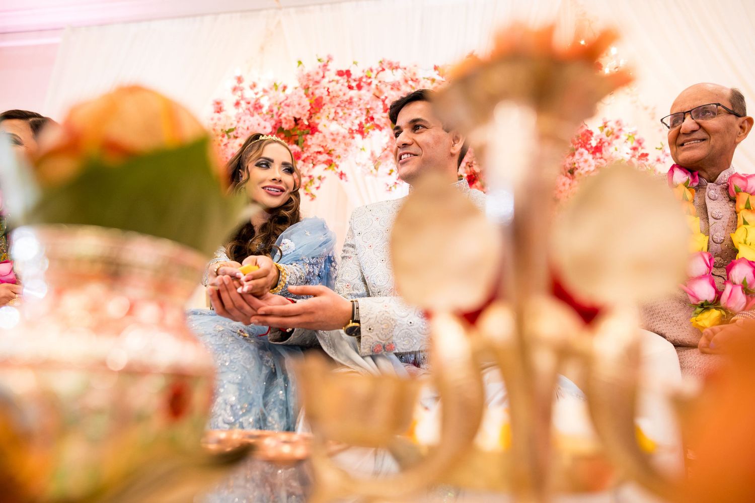 Couple in blue outfits smiling during a vibrant Indian wedding ceremony with floral decorations and golden ritual items.
