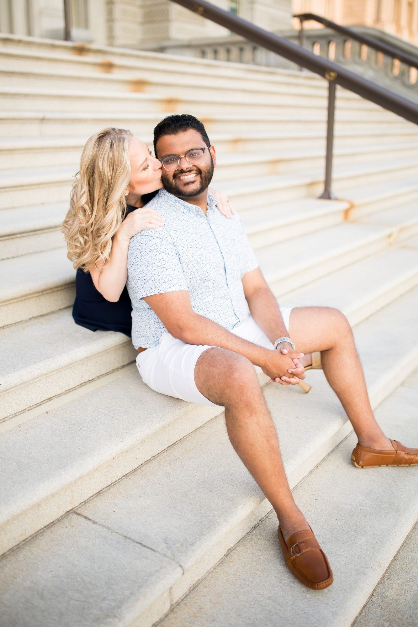 A couple sits on stone steps, woman kissing man's cheek as he smiles, wearing casual summer outfits.