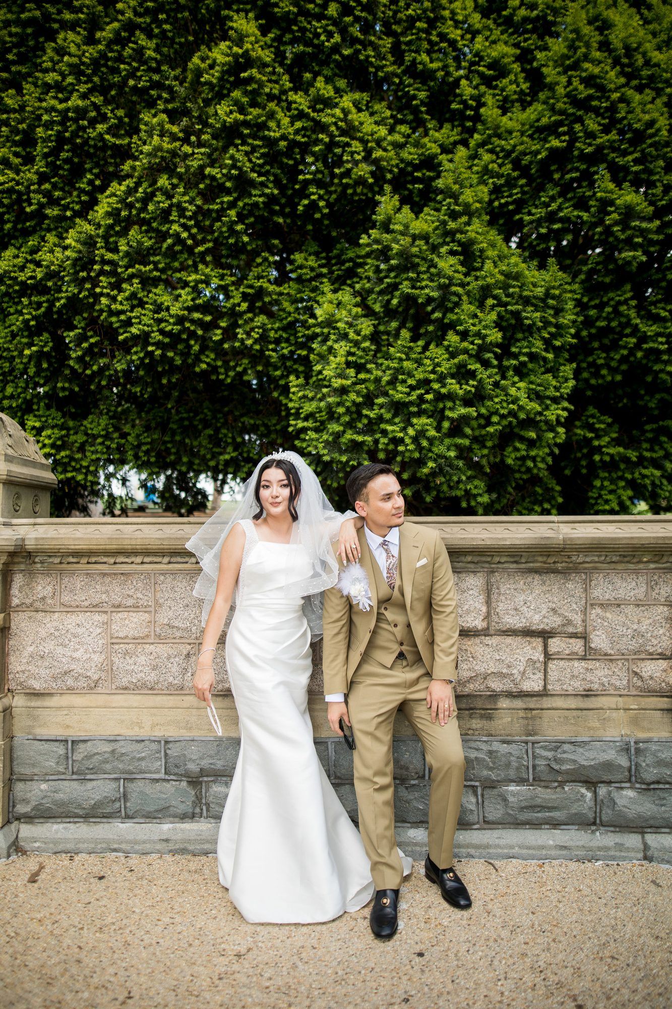 Bride in white gown and veil stands with groom in tan suit against stone wall with lush green trees.