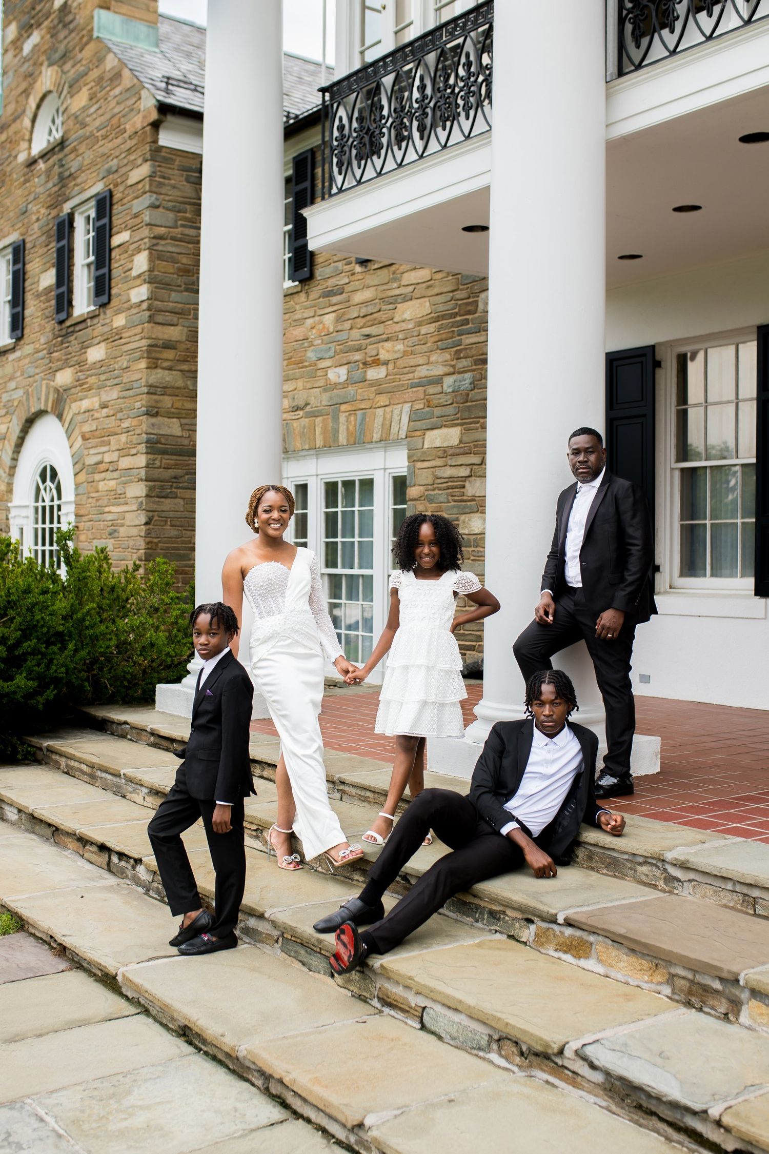 A joyful Black family poses on stone steps outside an elegant estate, with bride in white gown and others in formal attire.