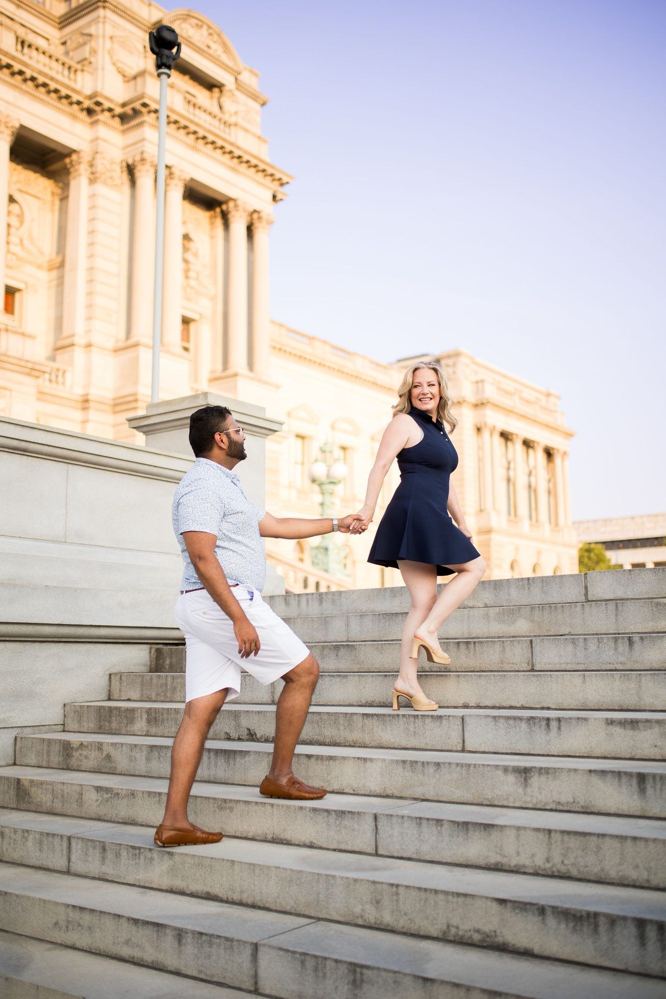 Couple holding hands on grand marble steps outside a neoclassical building at golden hour.