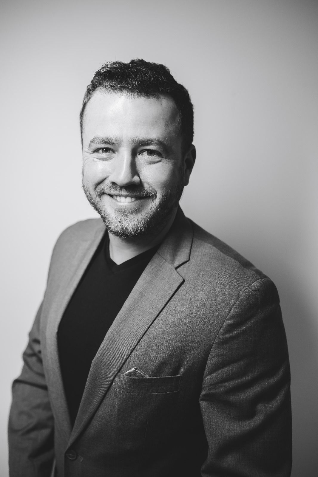 Smiling man in gray blazer and black shirt poses for professional black and white portrait photo.