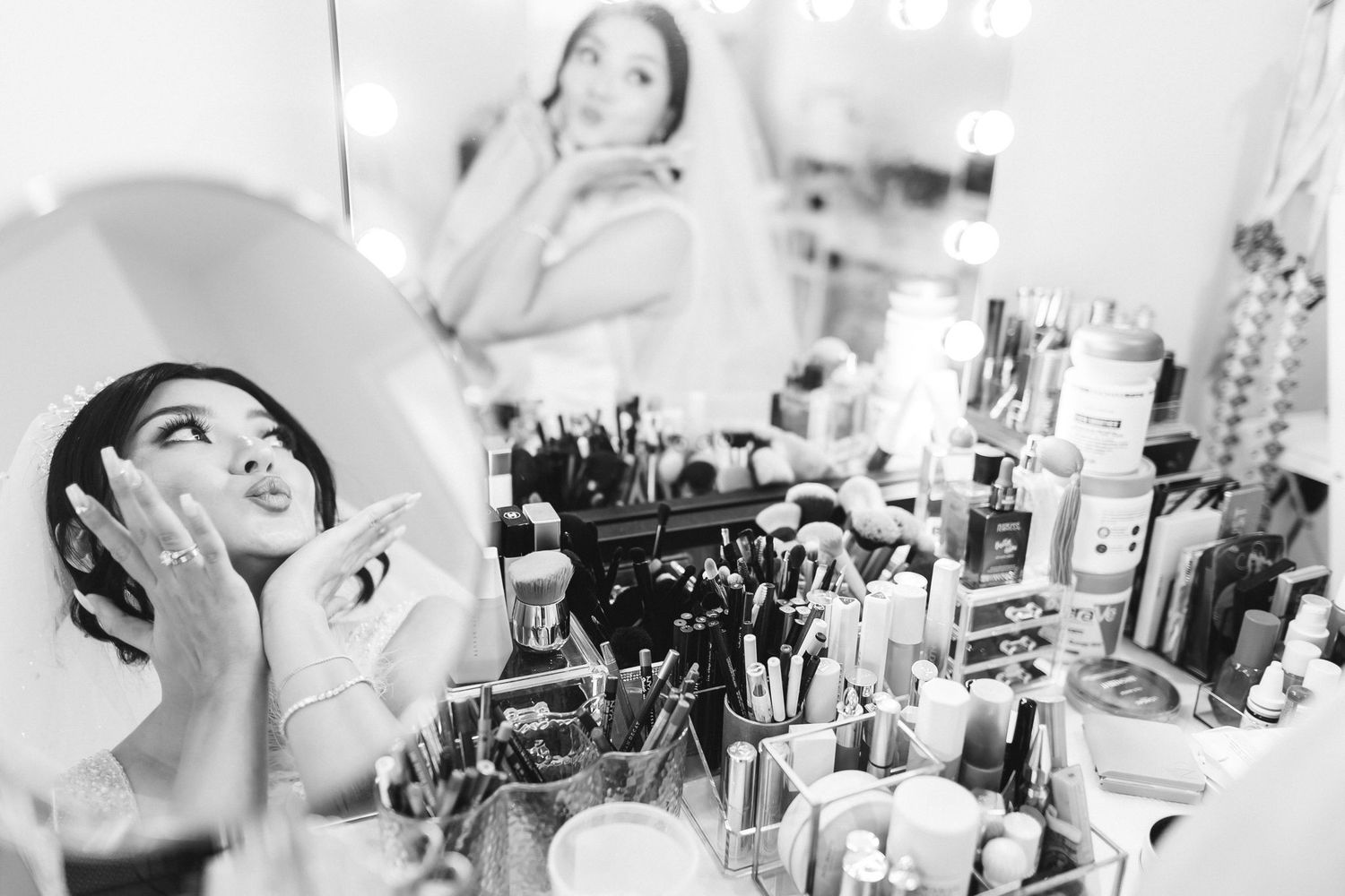 Black and white photo of a woman applying makeup at a vanity mirror surrounded by beauty products and brushes.