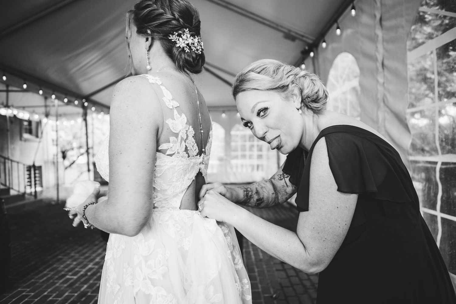 Black and white photo of a bridesmaid helping a bride with her wedding dress at an outdoor wedding reception.
