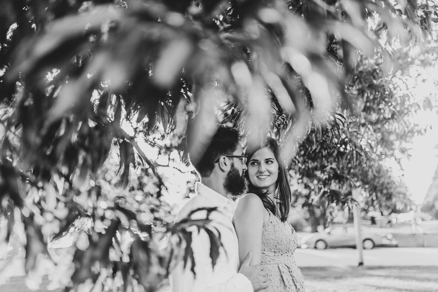 Black and white photo of a smiling couple framed by lush tree branches in an outdoor park setting.