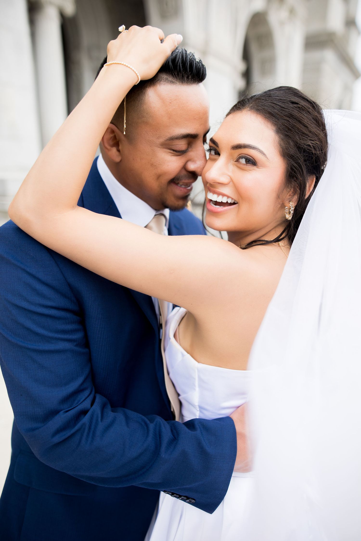 Joyful bride in white veil embraces groom in blue suit, sharing a romantic moment outside of the Library of Congress in Washington, DC.