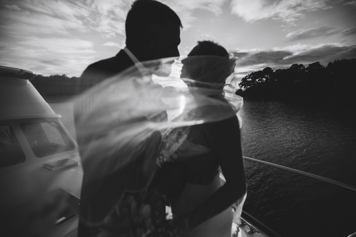 Black and white wedding photo of couple silhouetted against sunset sky near water, with dreamy double exposure effect.