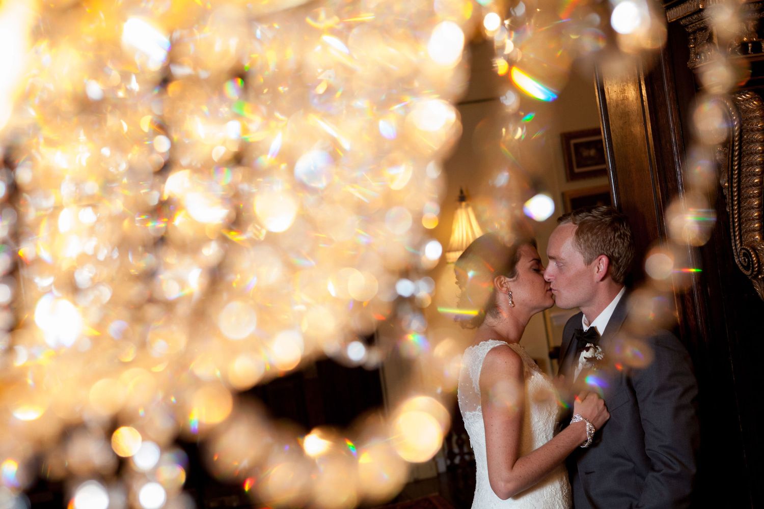 Couple sharing a romantic kiss surrounded by sparkling chandelier bokeh lights at an elegant wedding reception.