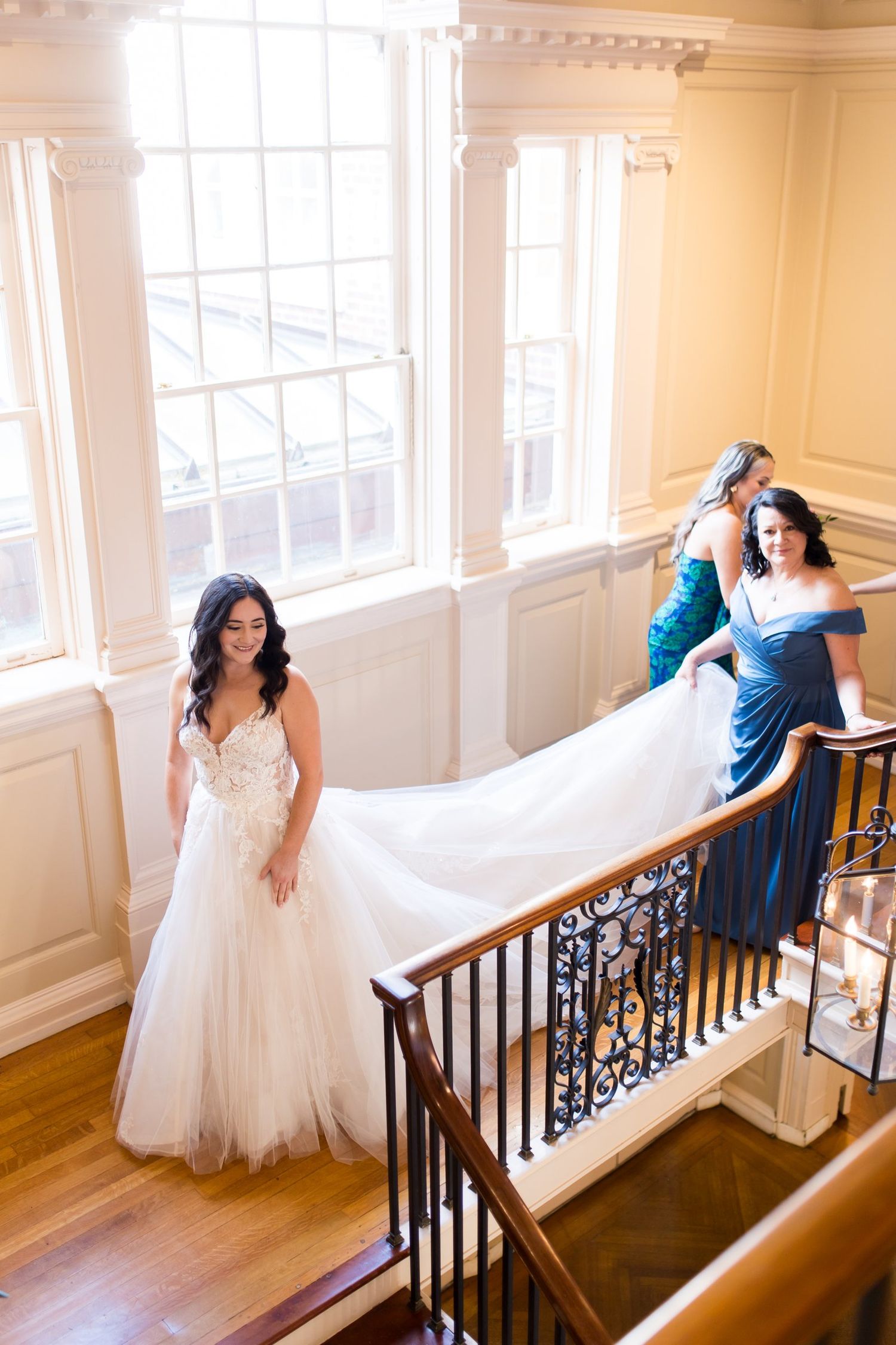 Bride in white lace gown descends grand staircase while bridesmaids in blue dresses hold her train.
