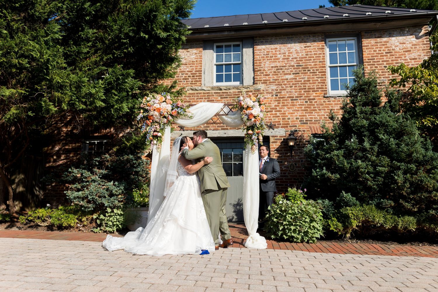 Bride and groom share a kiss under a floral arch at an outdoor wedding ceremony beside a brick building.