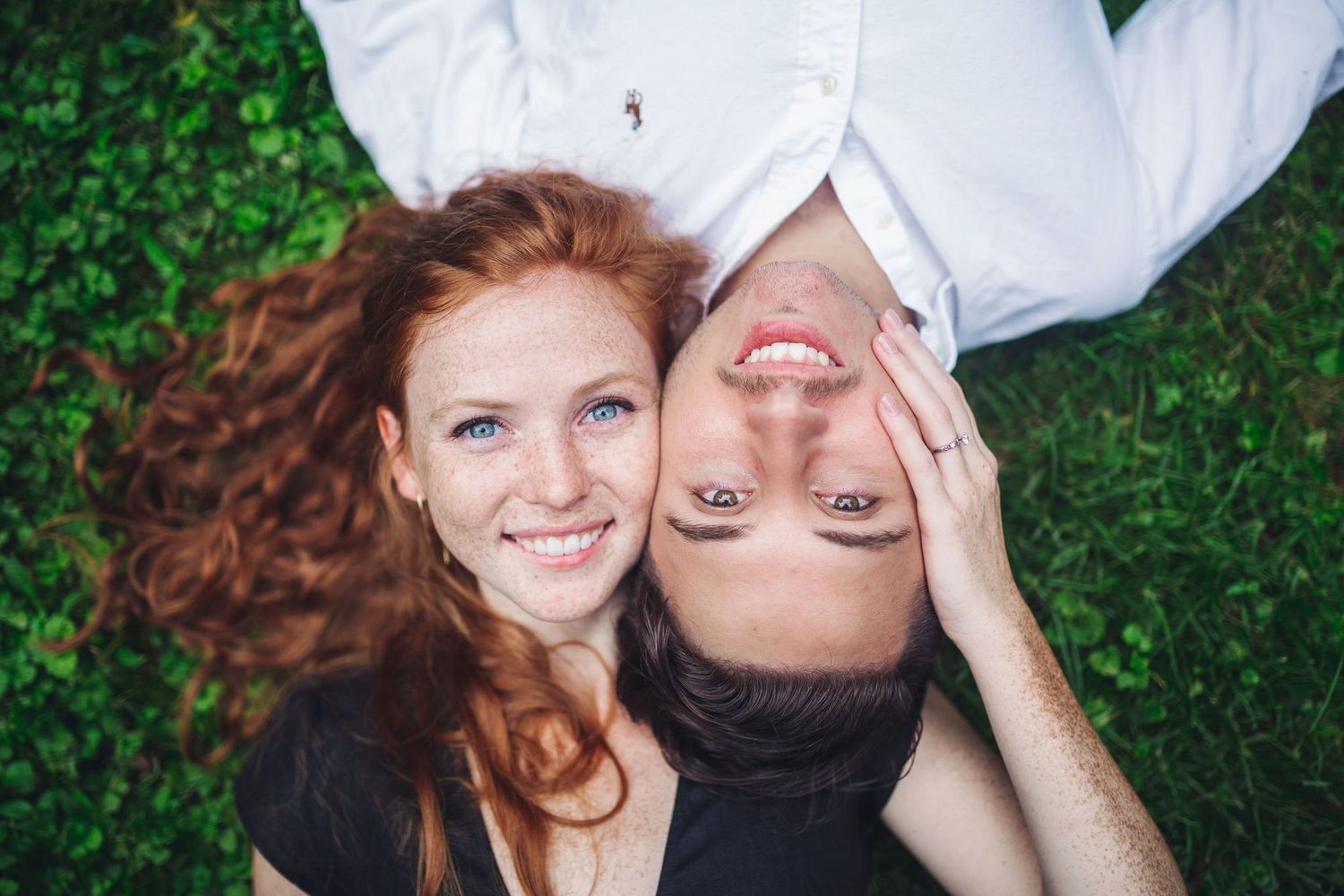 Two smiling friends lying on green grass, one with red curly hair and blue eyes, the other with dark hair.
