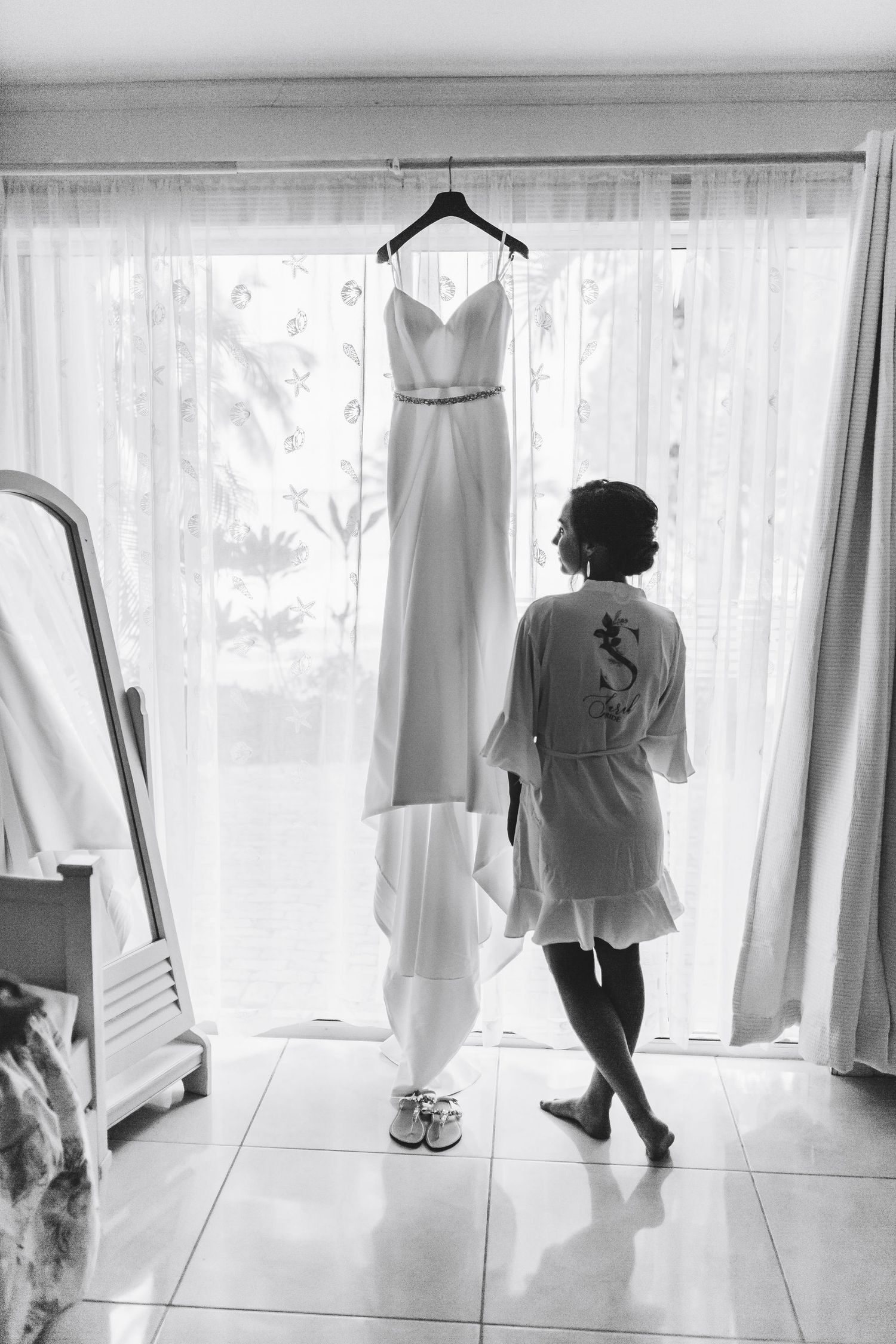 Bride in robe admiring elegant wedding gown hanging by window in black and white photo.