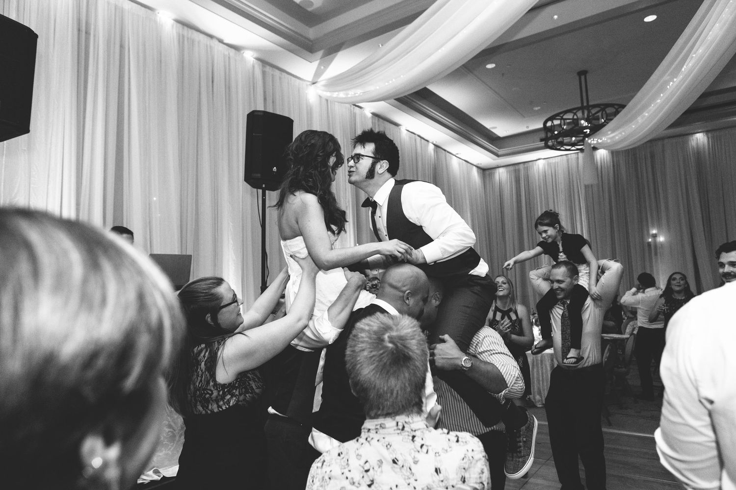 Black and white photo of bride and groom kissing while being lifted by guests on wedding reception dance floor.
