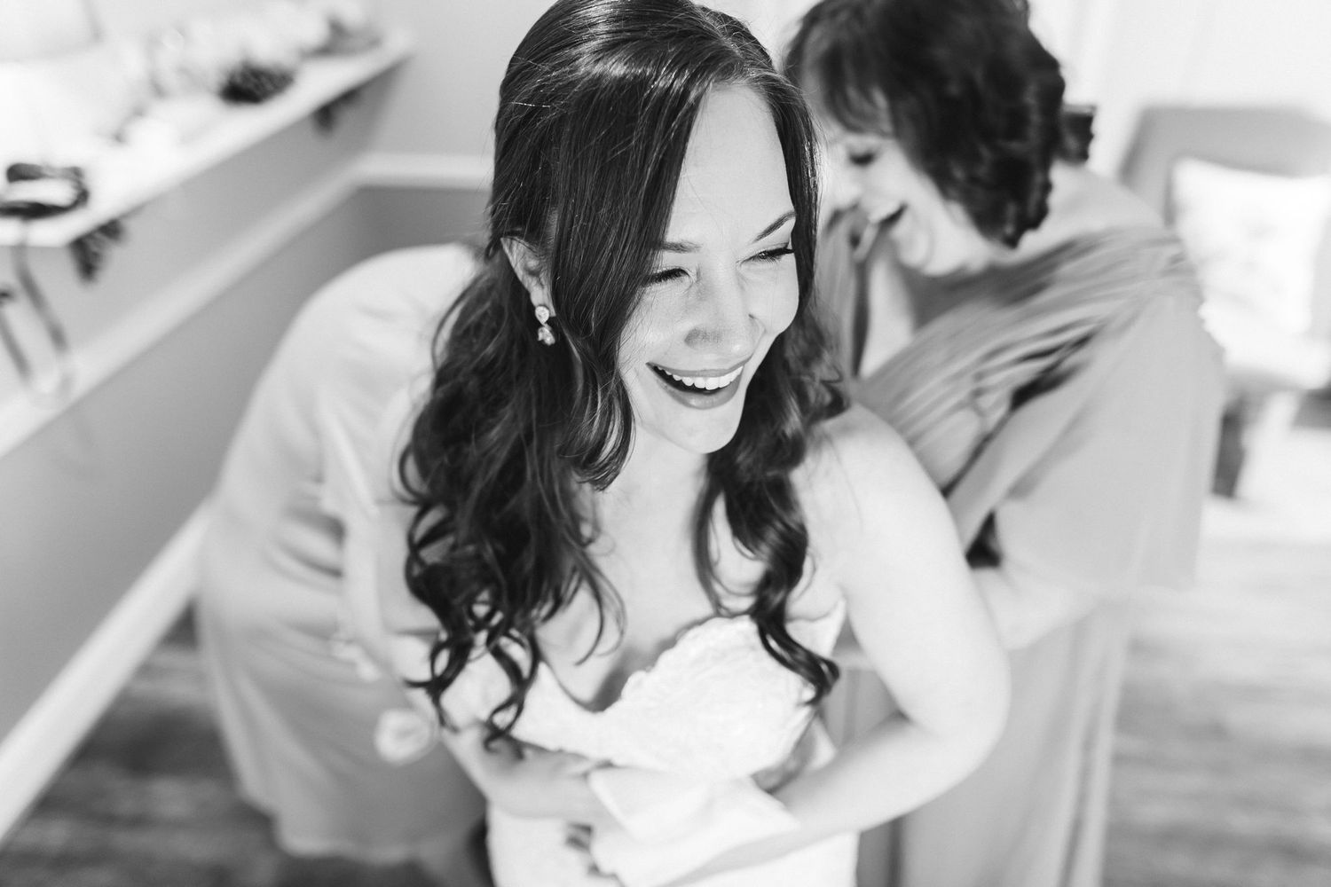 Bride laughing joyfully in strapless wedding gown while bridesmaid helps with dress in black and white photo.