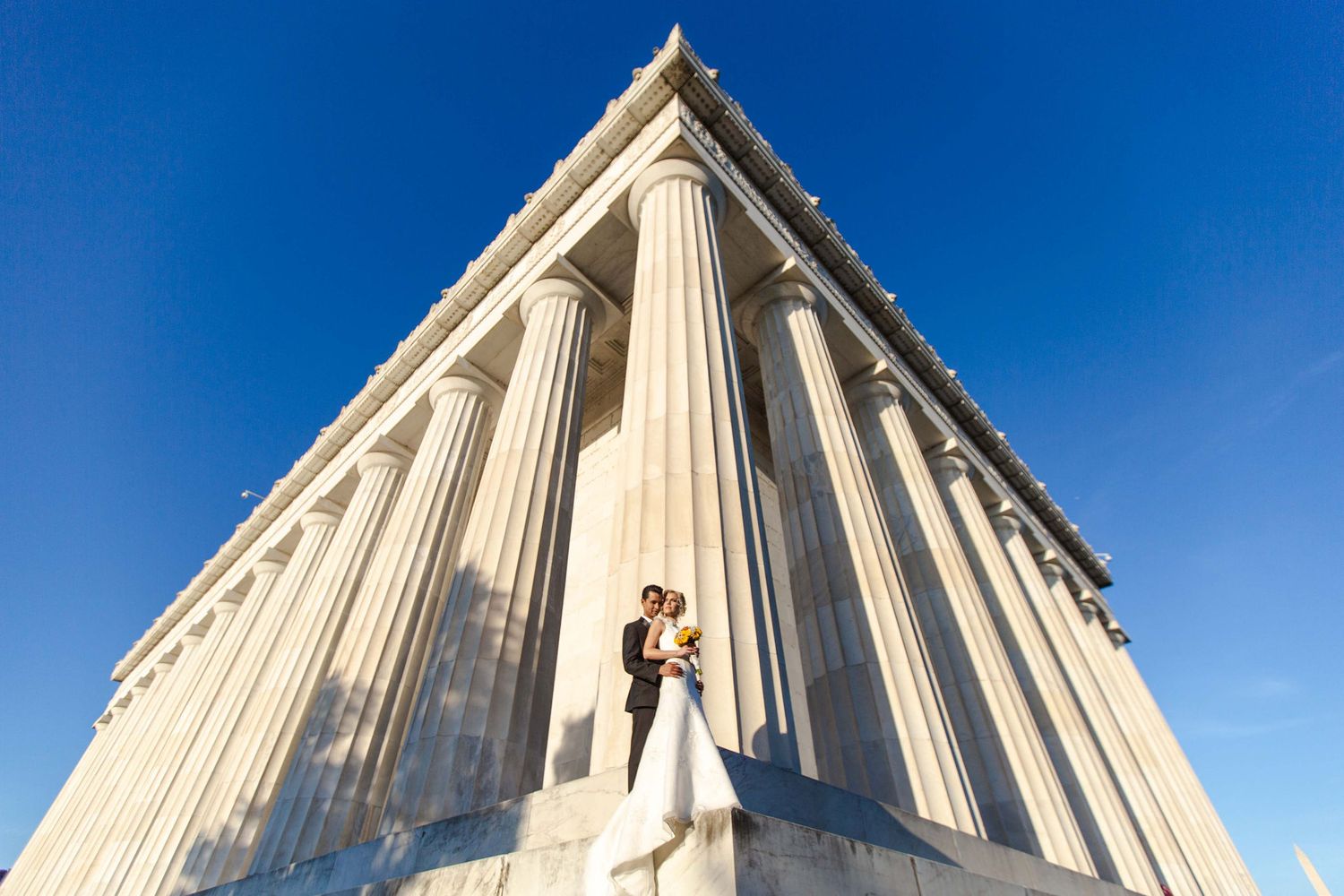 Bride and groom embrace among towering white columns of a neoclassical building under a vivid blue sky.