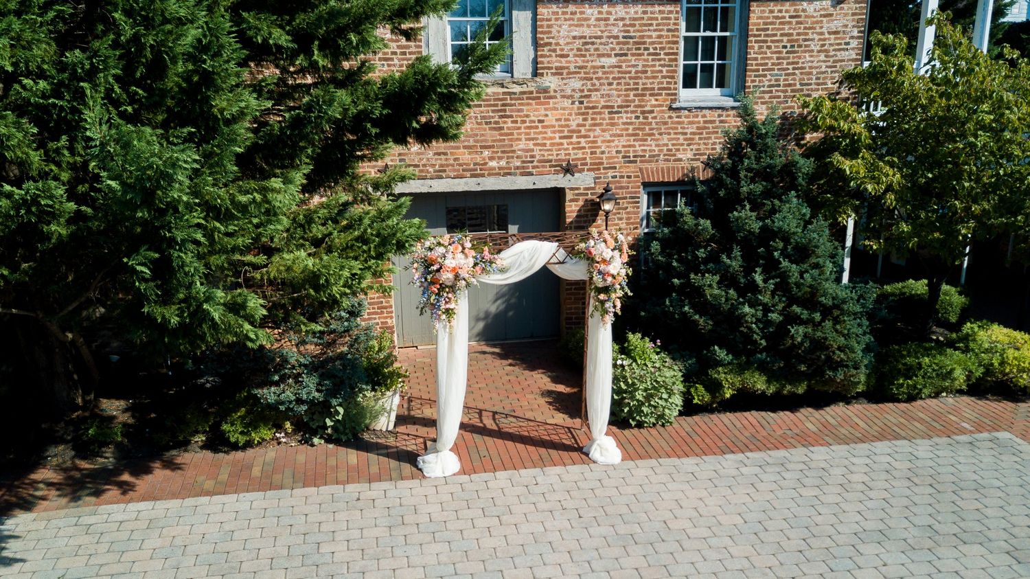 Elegant floral wedding arch with white columns and pink blooms at brick building entrance on herringbone brick path.