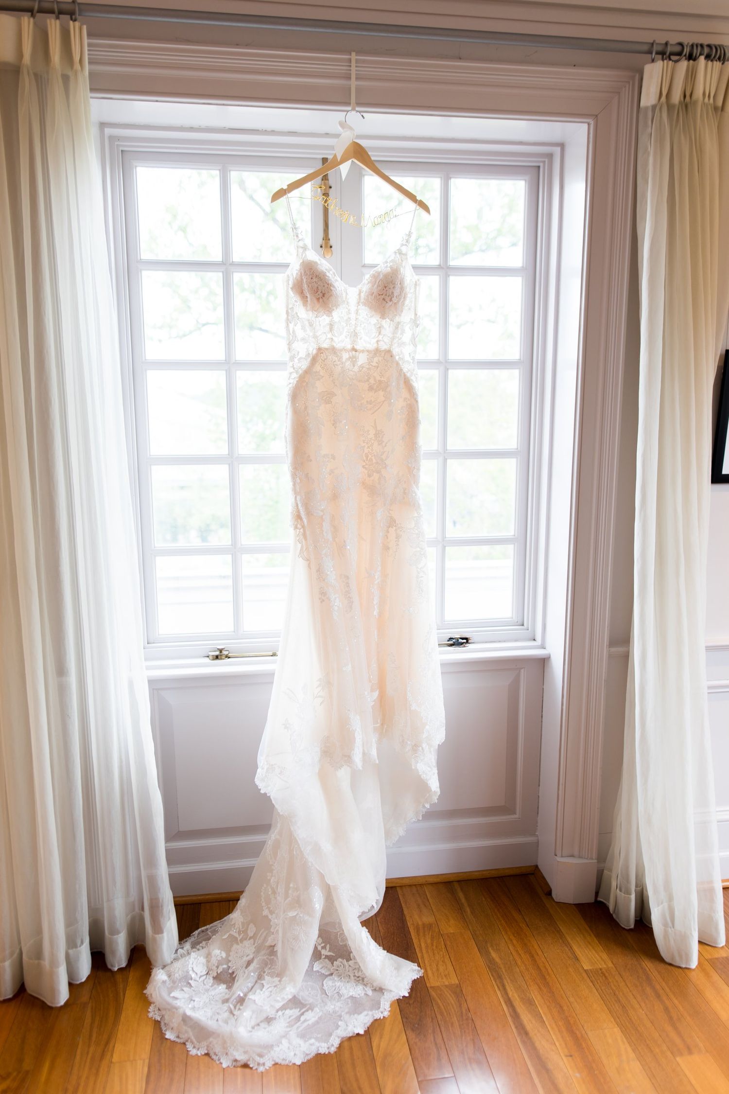 Elegant lace wedding dress hanging on a wooden hanger in front of a bright white window with sheer curtains.