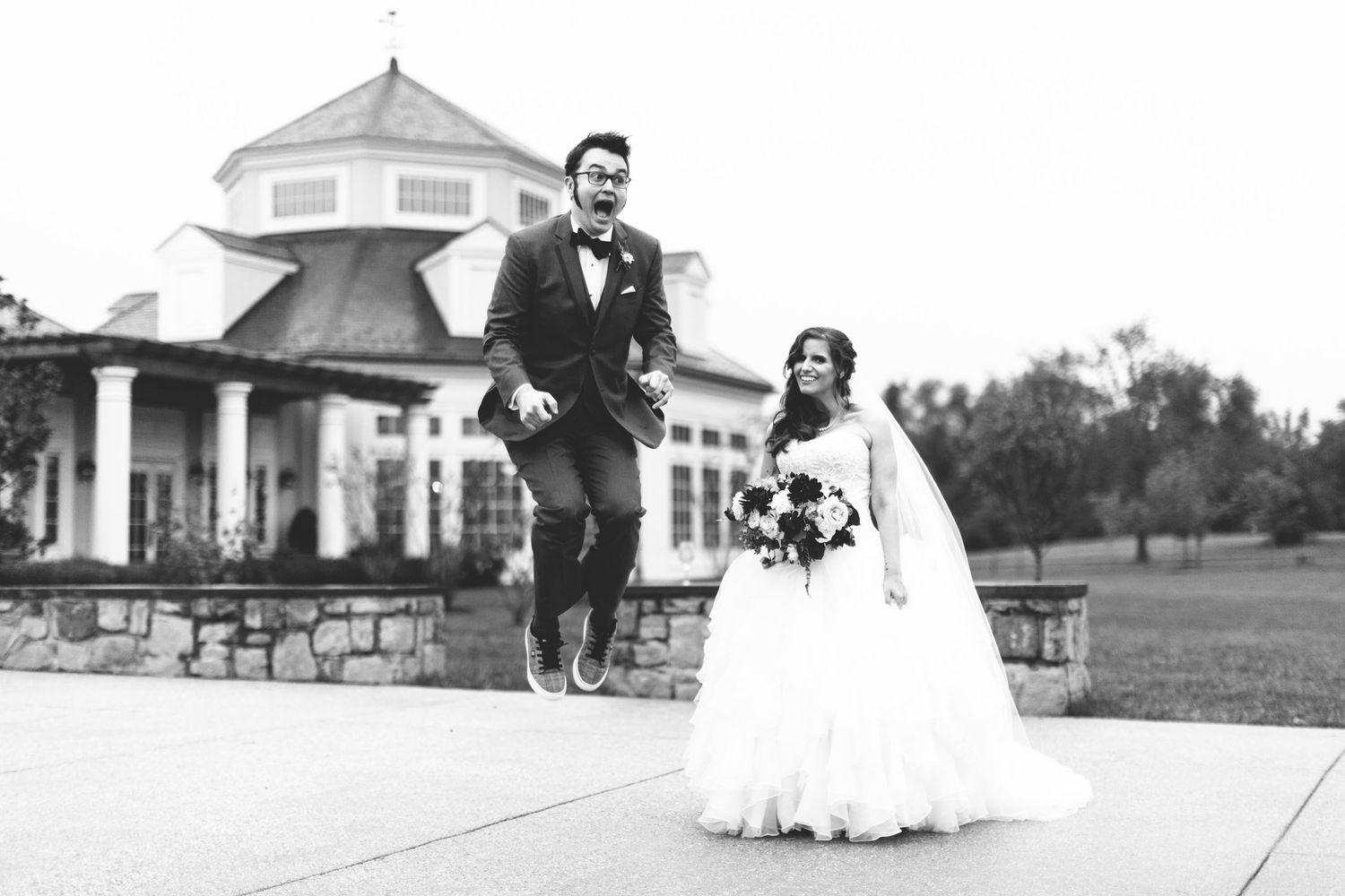 Black and white photo of a joyful bride and groom jumping outside an elegant wedding venue.