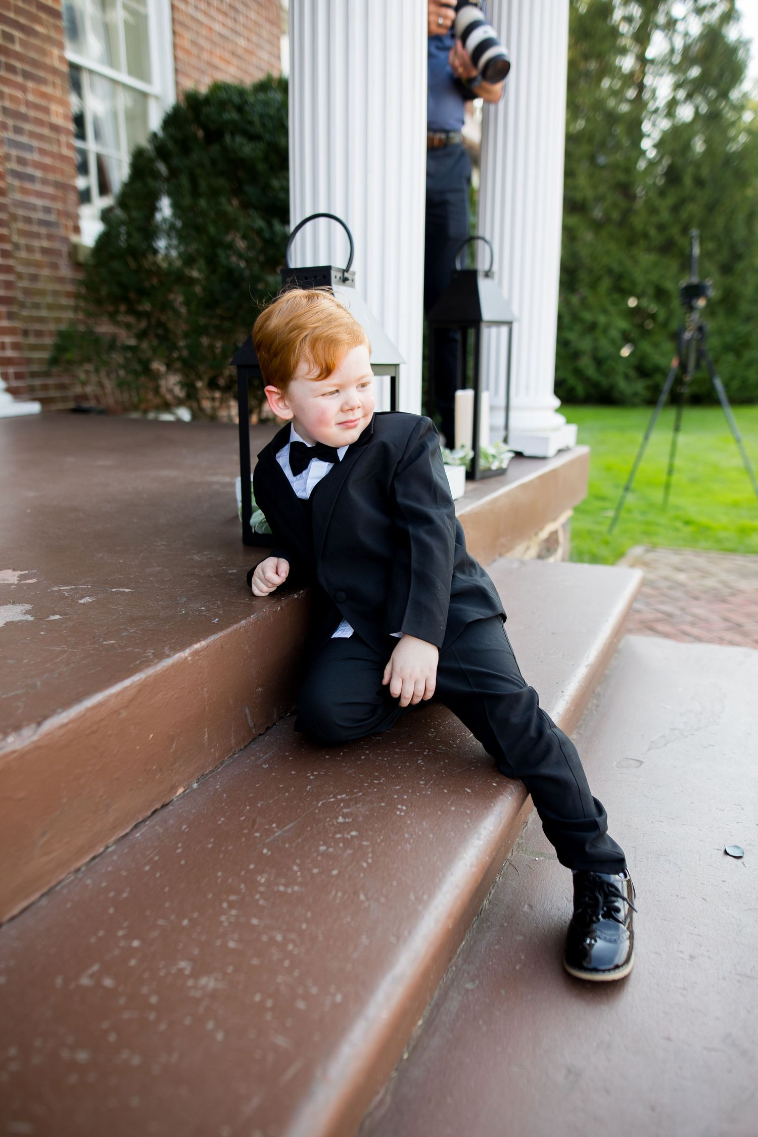 Adorable redhead boy in black tuxedo and bow tie sitting on steps of elegant building.