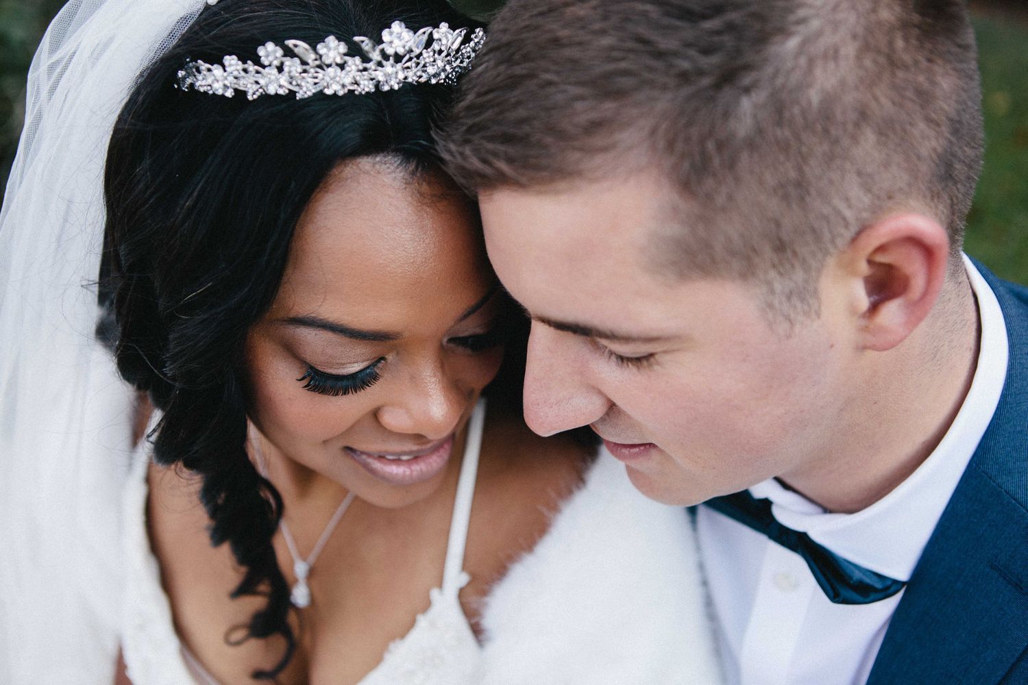 Interracial couple in wedding attire, bride wearing tiara and veil, groom in blue suit, sharing a romantic moment.
