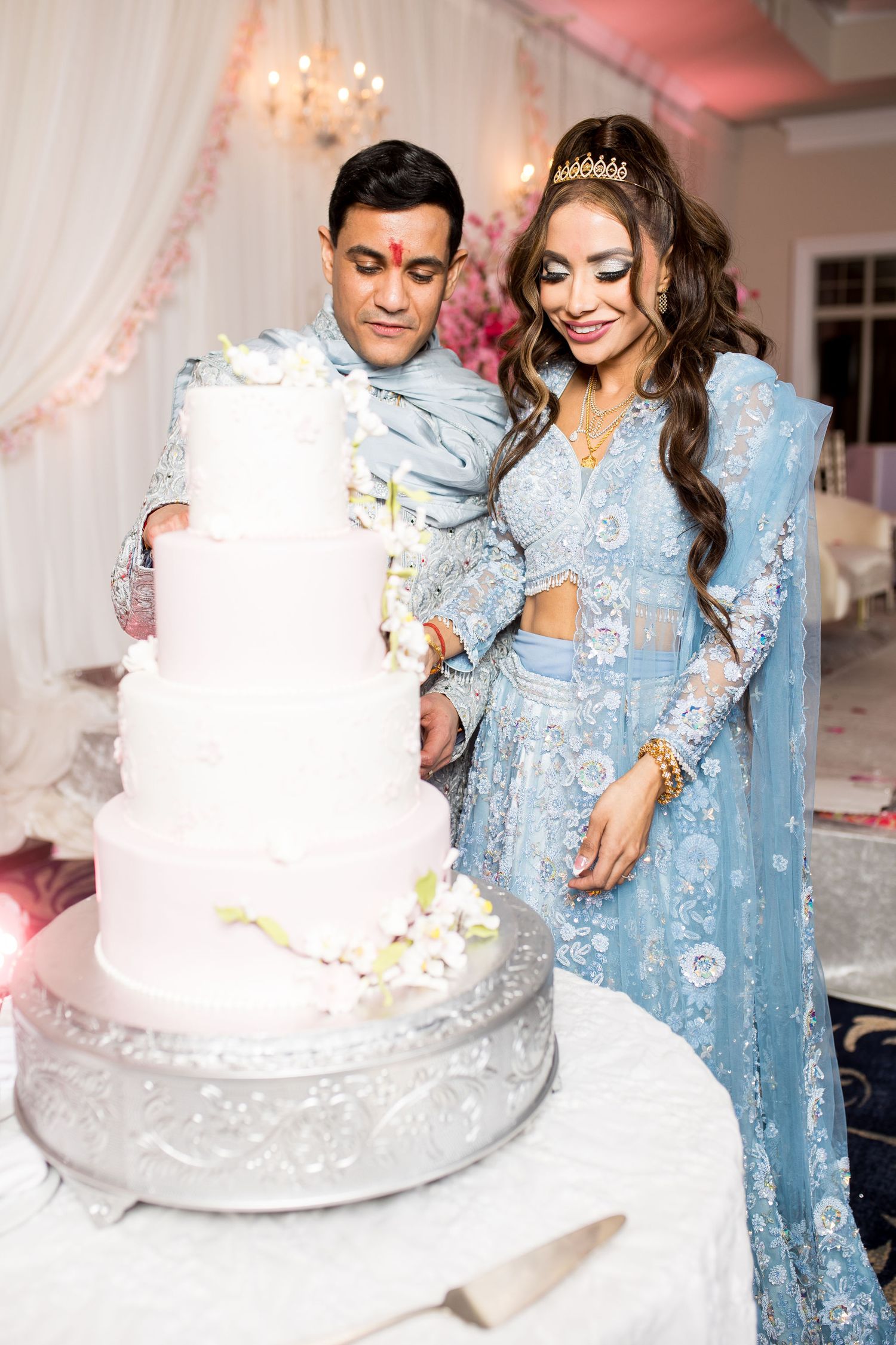 Bride and groom in blue traditional attire cutting a four-tier white wedding cake at an elegant reception.