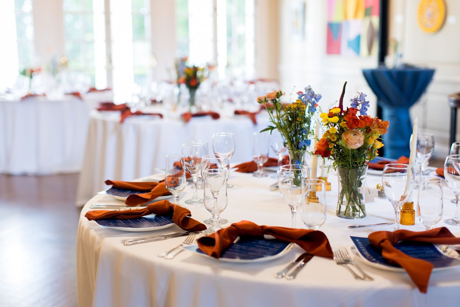 Elegant wedding reception table set with orange napkins, blue chargers, wildflower centerpieces, and glassware in a bright venue.