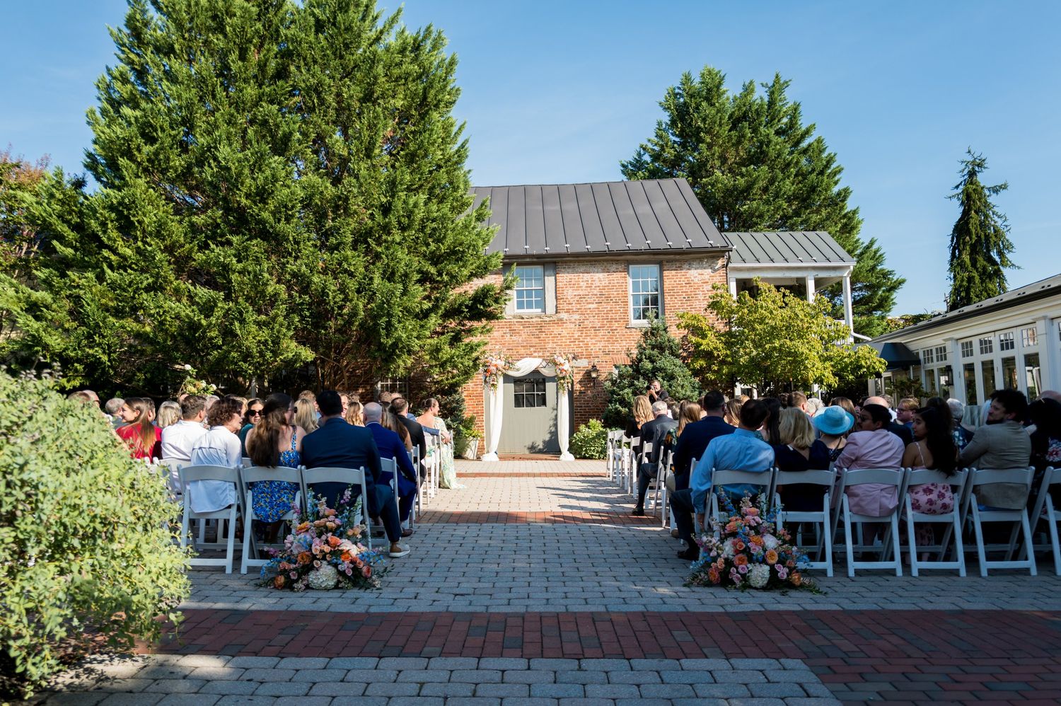 Outdoor wedding ceremony on brick patio with white chairs, lush trees, and elegant brick building backdrop on sunny day.