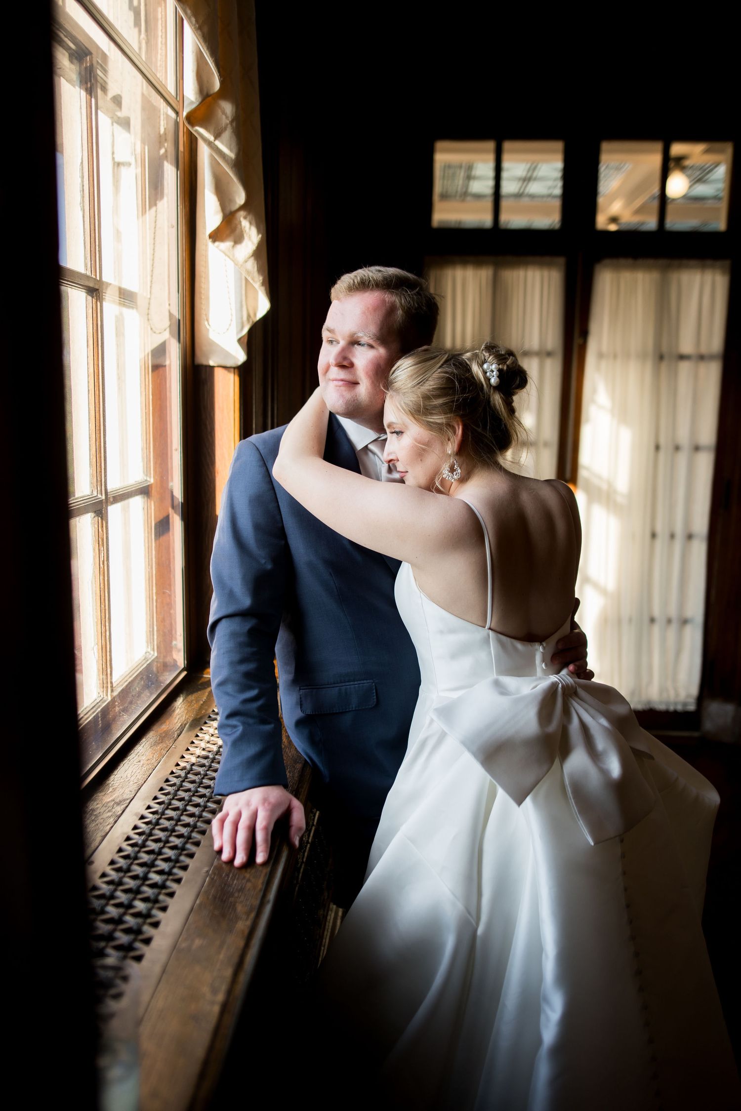 Bride in white gown with bow embraces groom in navy suit by sunlit windows in elegant wedding portrait.