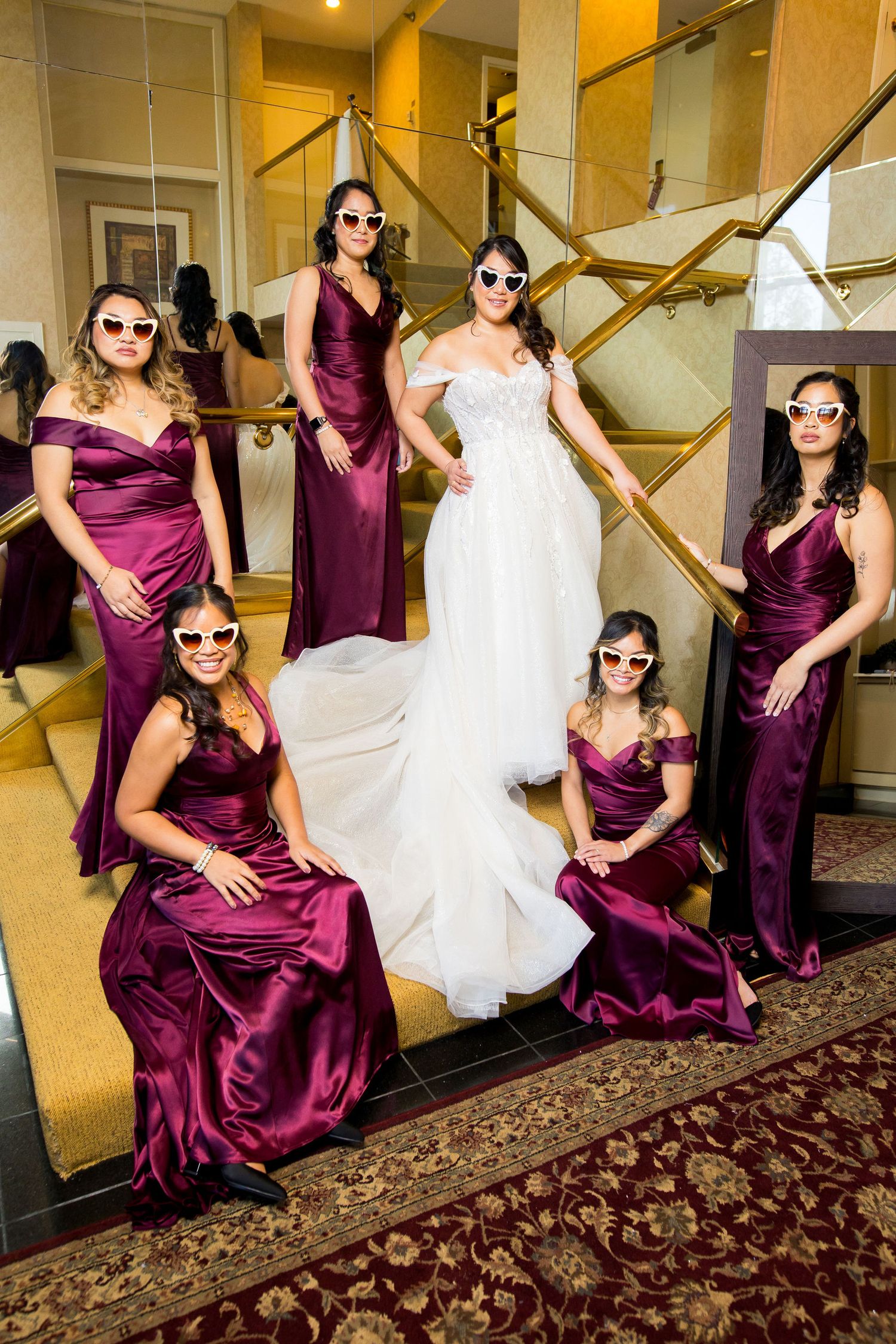 Bride in white gown poses with bridesmaids in burgundy satin dresses and white heart sunglasses on elegant staircase.