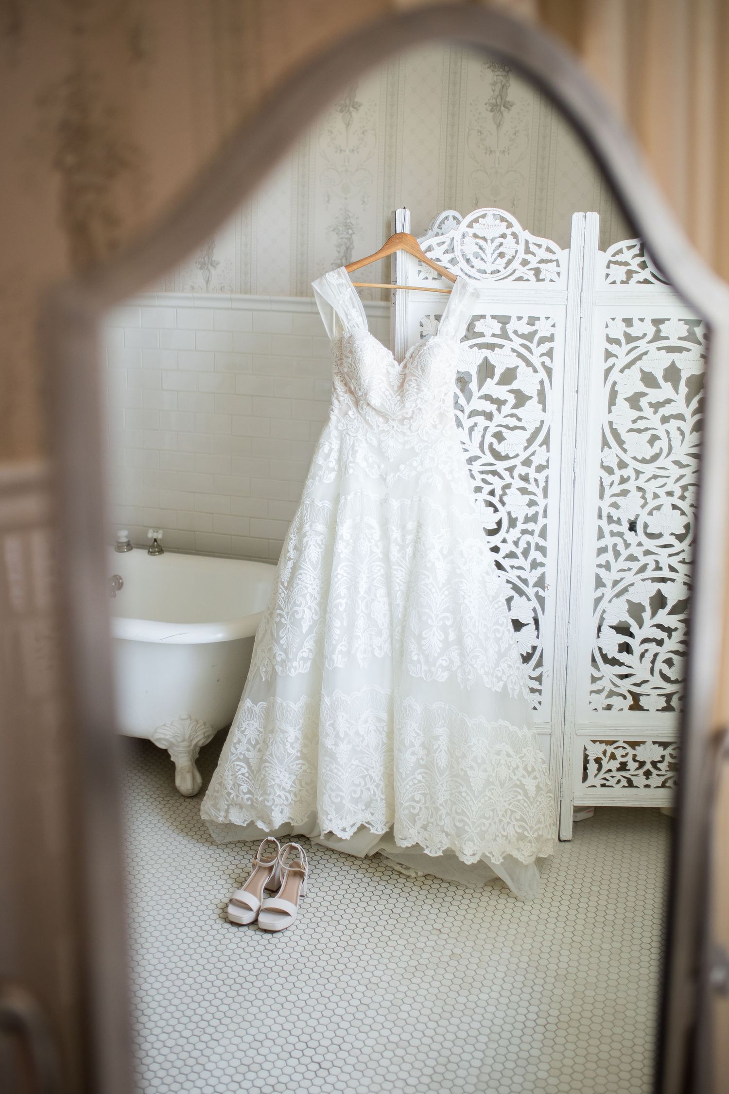 White lace wedding dress hanging on ornate screen reflected in vintage mirror with bridal heels on tiled floor.