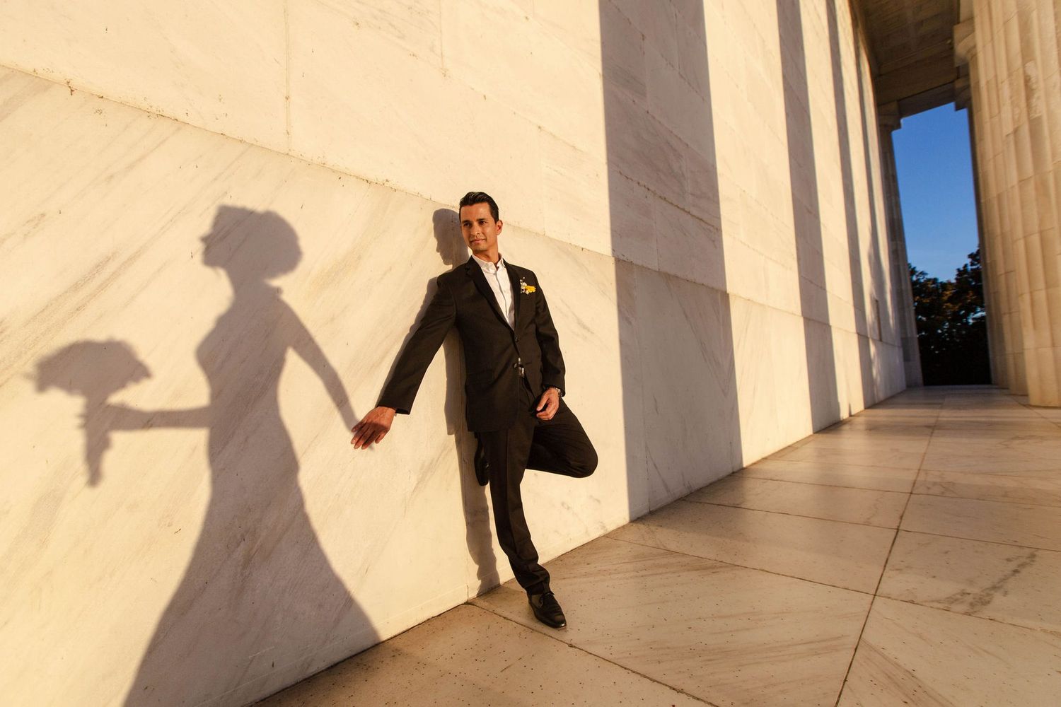 Groom in black suit leans against marble column at Lincoln Memorial, with bride's shadow holding bouquet on wall.
