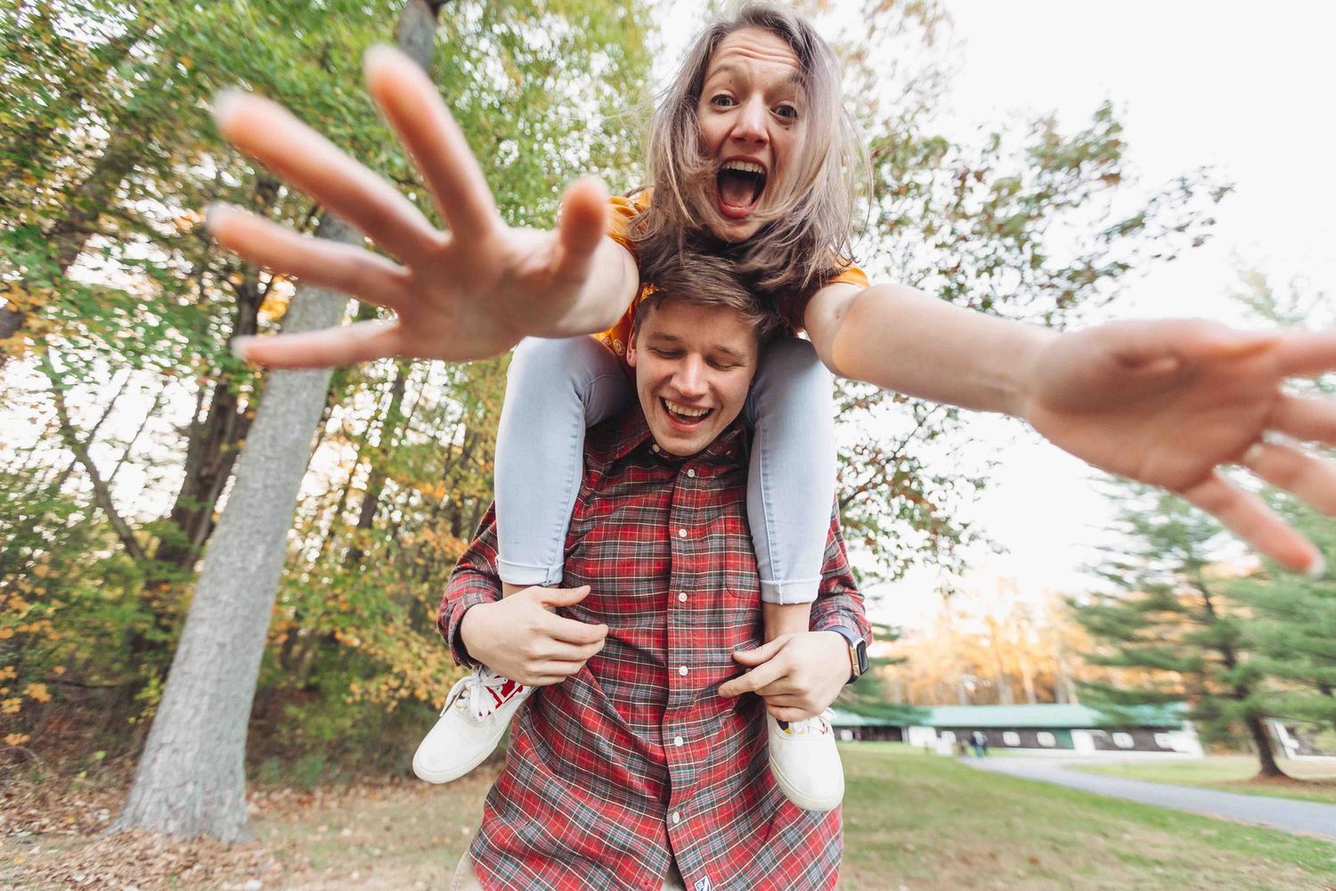 Two playful women enjoy a piggyback ride outdoors, laughing and reaching toward the camera in a colorful autumn park.