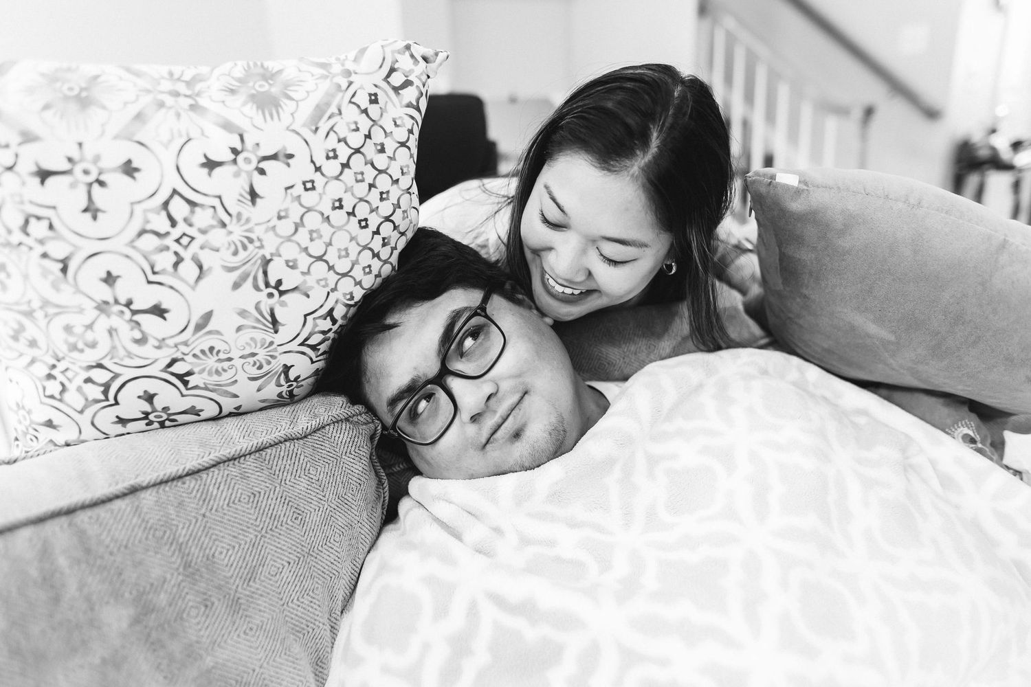 Black and white photo of a couple relaxing on a couch, woman smiling down at man wearing glasses under a blanket.
