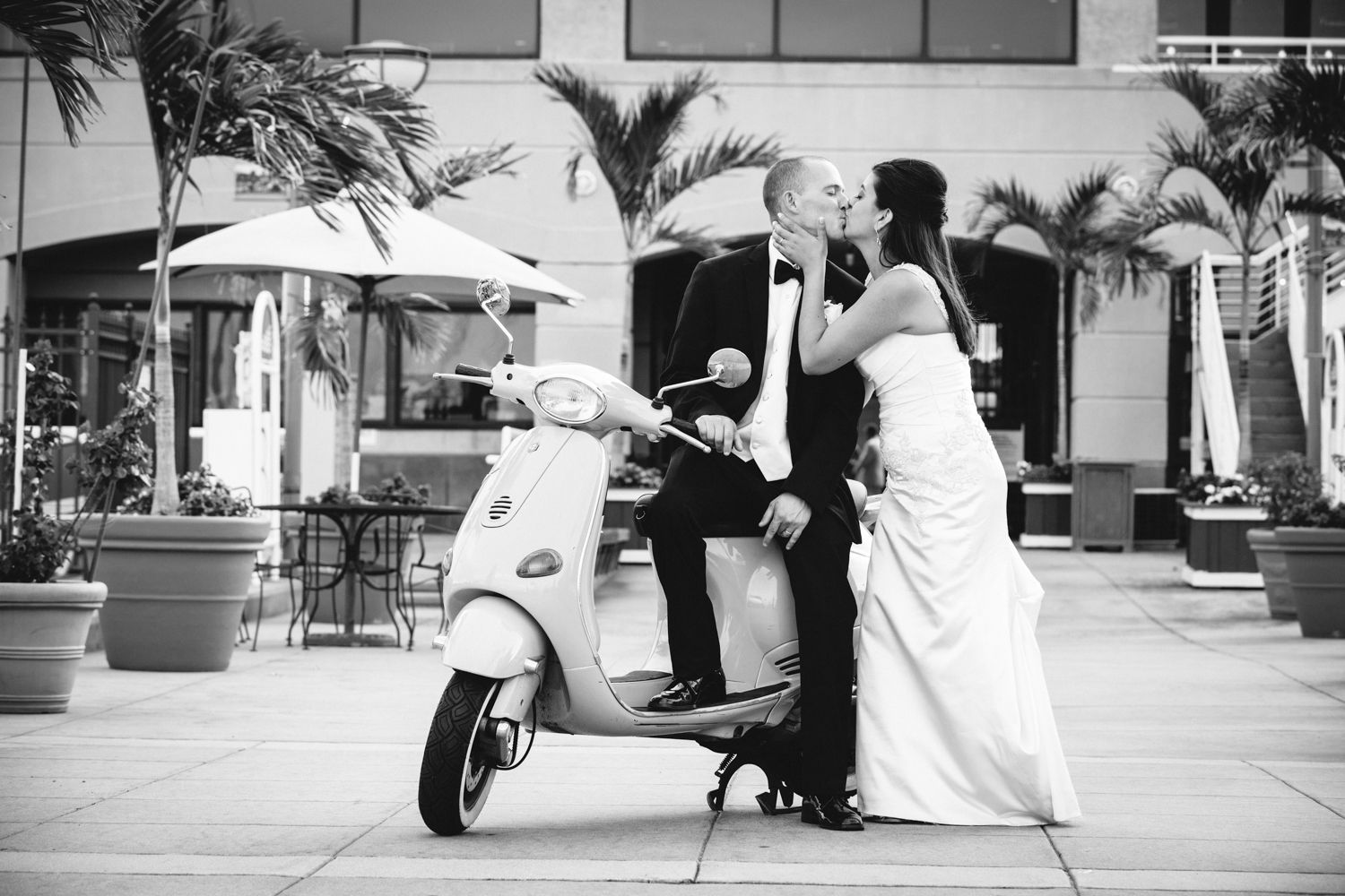Bride and groom kissing on a white Vespa scooter in an outdoor plaza with palm trees in black and white.